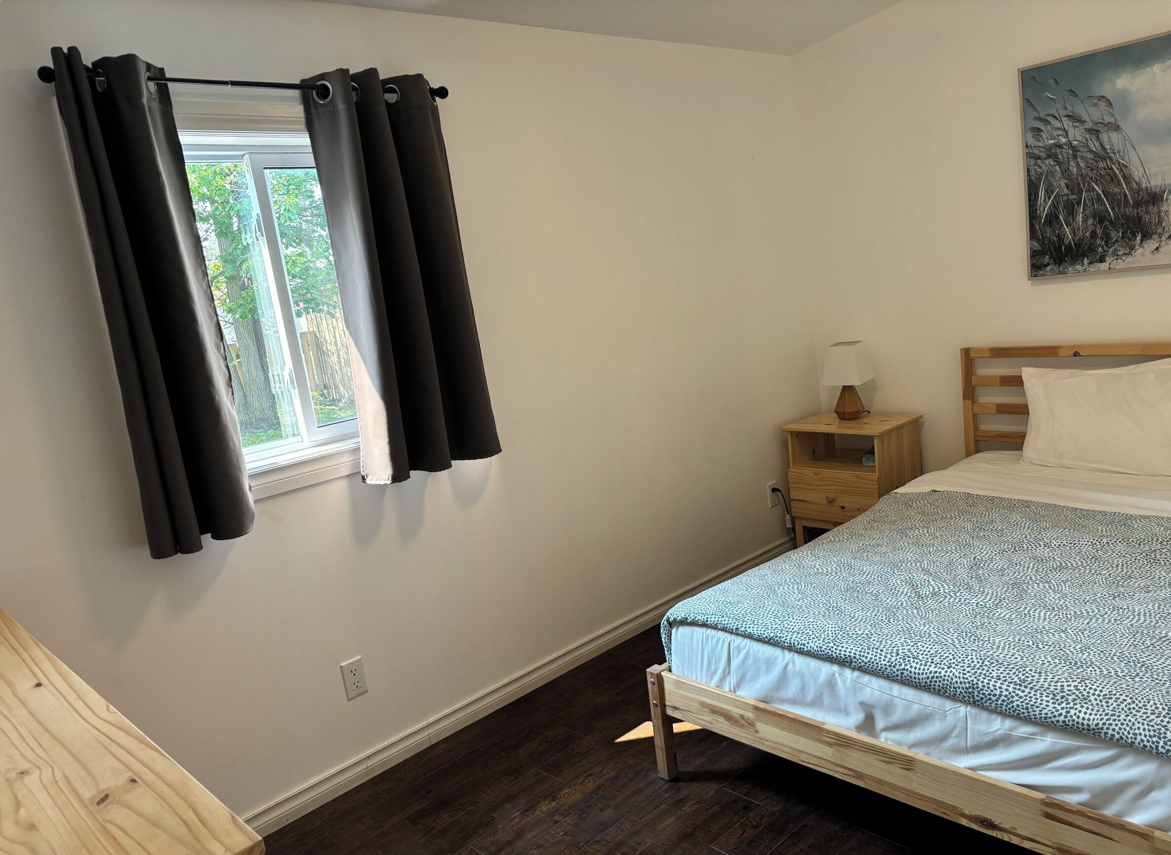 A bedroom with a bed, a nightstand, a lamp, a window with curtains, and a painting on the wall.
