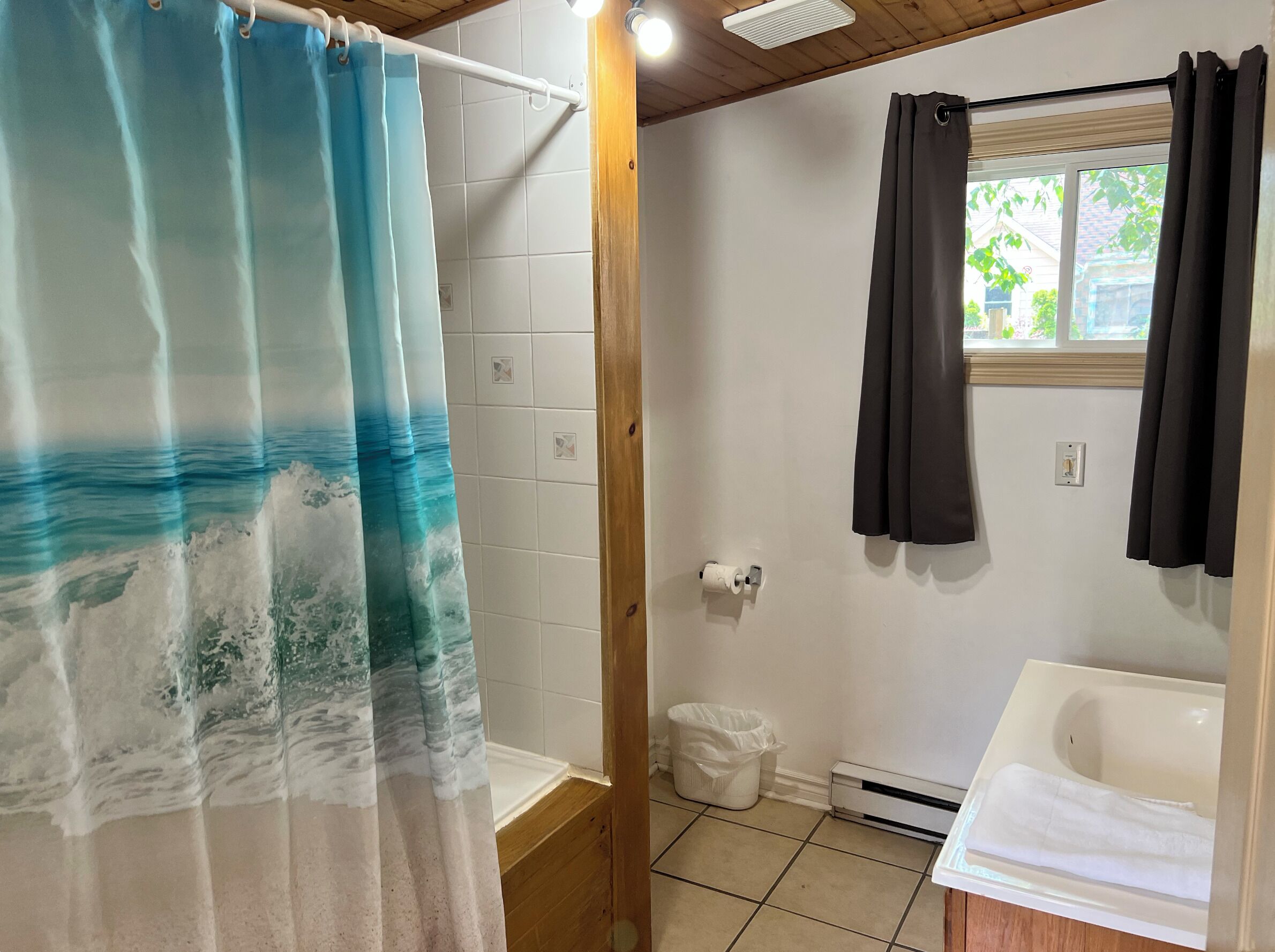 A bathroom with a shower curtain depicting ocean waves, a white bathtub, a window with dark curtains, and a white trash bin.