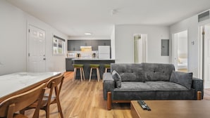 A modern living room with a grey sofa, wooden dining table, and chairs. A kitchen area with a white fridge and grey cabinets is visible in the background. - Saga Resort (Wasaga Beach)