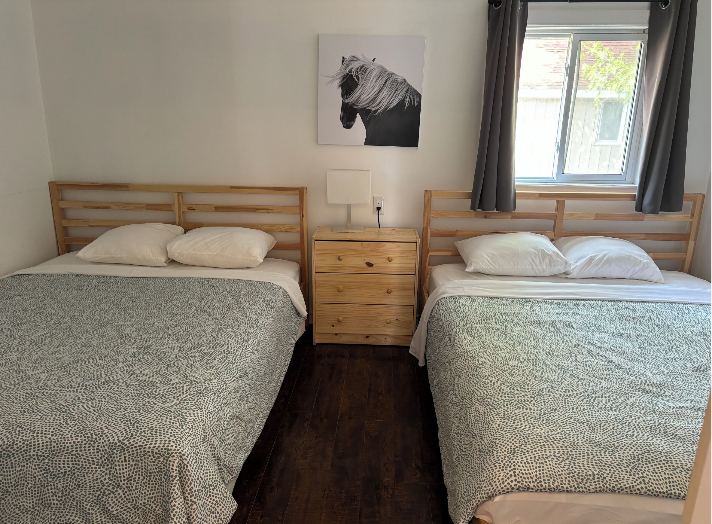 Ein Doppelzimmer mit zwei Betten, einem Holzkopfstück, einem Nachttisch, einer Lampe und einem Vorhangfenster.