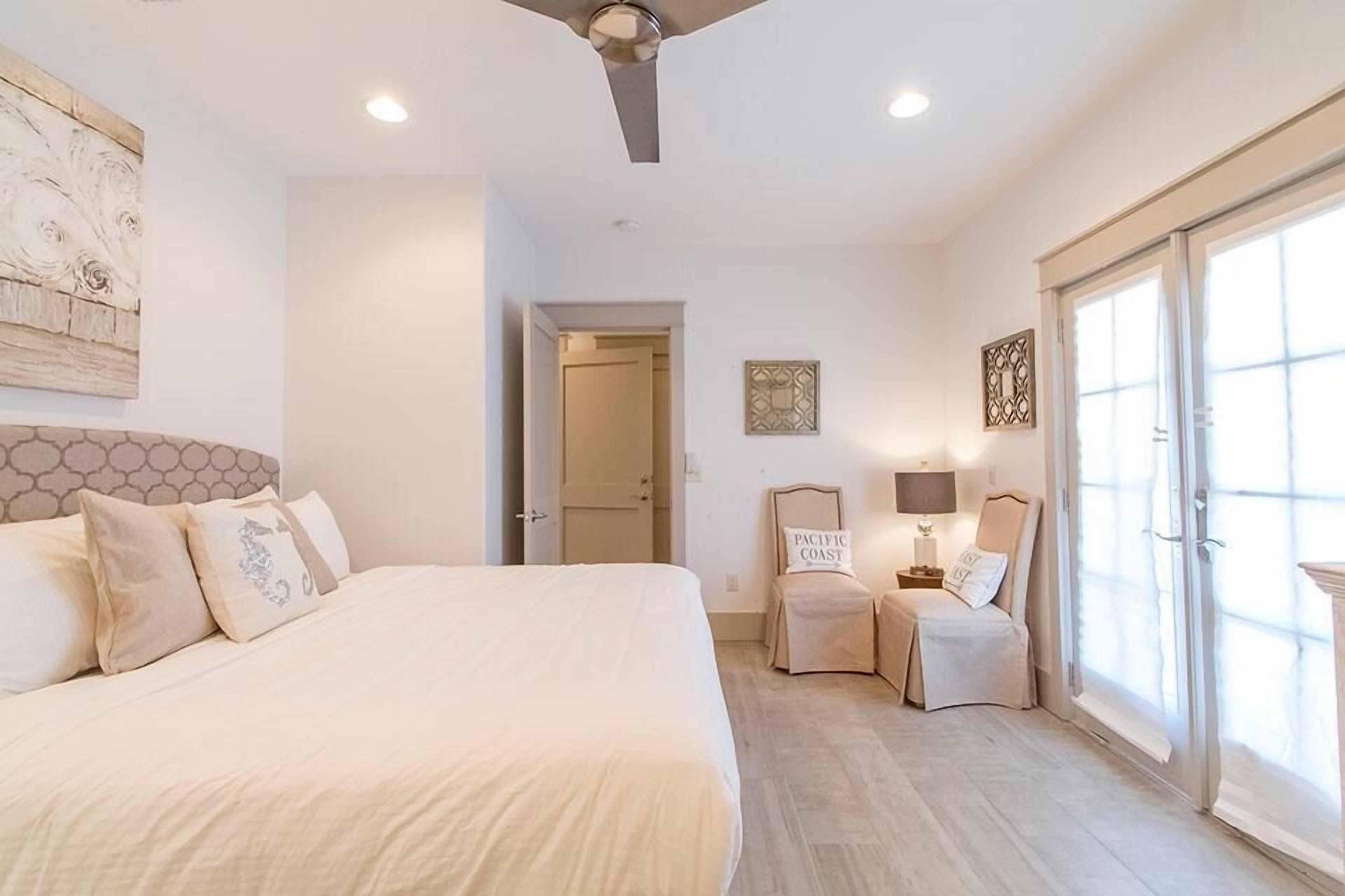 A bedroom with a large bed, a ceiling fan, a door leading to another room, and a window with a view.