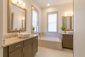 A modern bathroom with a large mirror, two sinks, a bathtub, and a window.