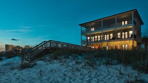 Un edificio de varios pisos frente a la playa, con una escalera que conduce a un balcón, rodeado de arena y vegetación.