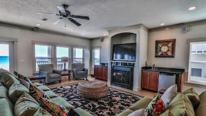 A spacious living room with a large green sofa, a round ottoman, and a fireplace. There are two windows with a view of the ocean. - Seaclusion 9 Bedroom Holiday Home by Five Star Properties (Miramar Beach)