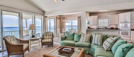A spacious living area with a green sofa, a wooden coffee table, and a dining table with chairs. There is a kitchen area with white cabinets and a large window overlooking the ocean.