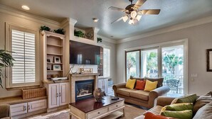 A living room with a fireplace, a ceiling fan, and a large window with a view of palm trees. - Serenity 4 Bedroom Holiday Home by Five Star Properties (Destin)