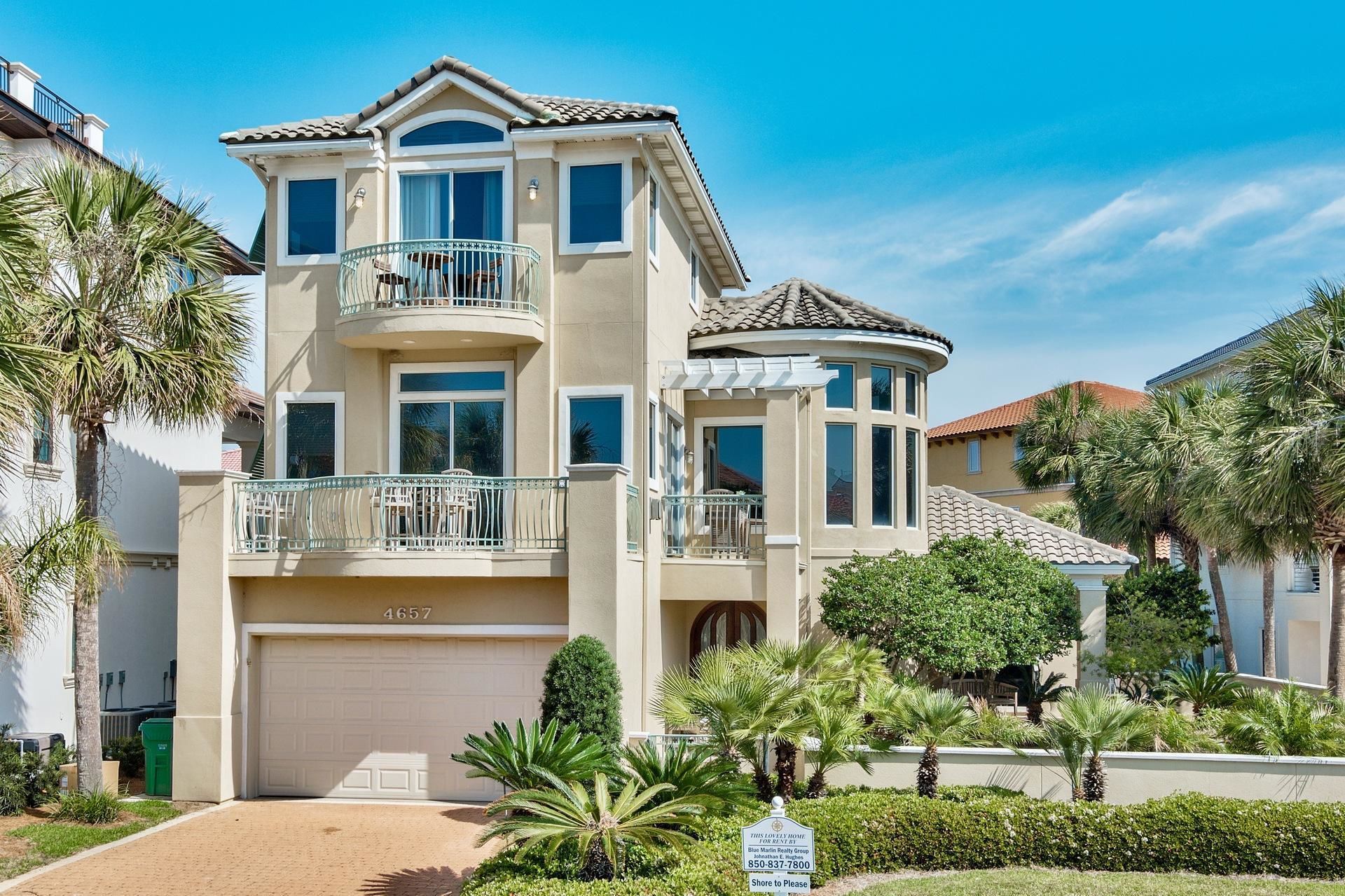 A two-story house with a garage, a balcony, and a well-maintained garden with palm trees.