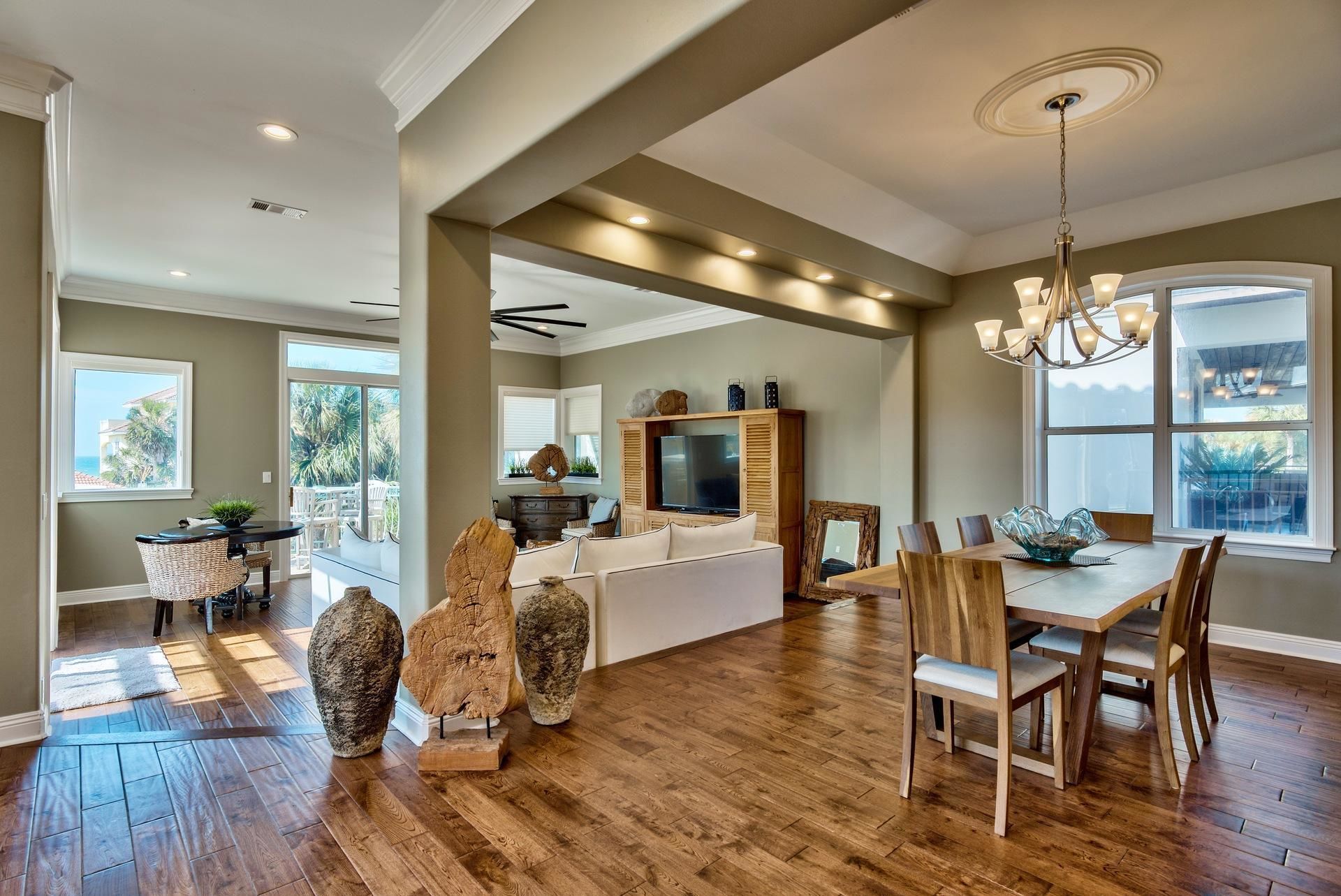 A spacious living area with a wooden dining table, a sofa, and a TV on a wooden cabinet. There are large windows providing natural light and a view of the outdoors.