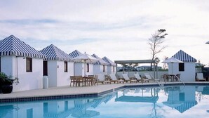Zona de piscina con múltiples sillas de descanso y sombrillas, una piscina cristalina que refleja el entorno, y una fila de edificios pequeños con techos a rayas.