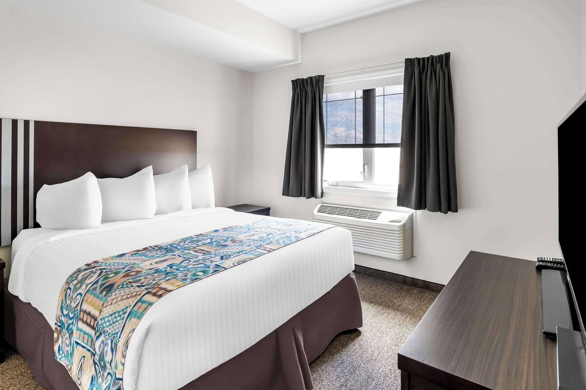 A hotel room with a large bed, a desk, a window with curtains, and a wall-mounted air conditioner.