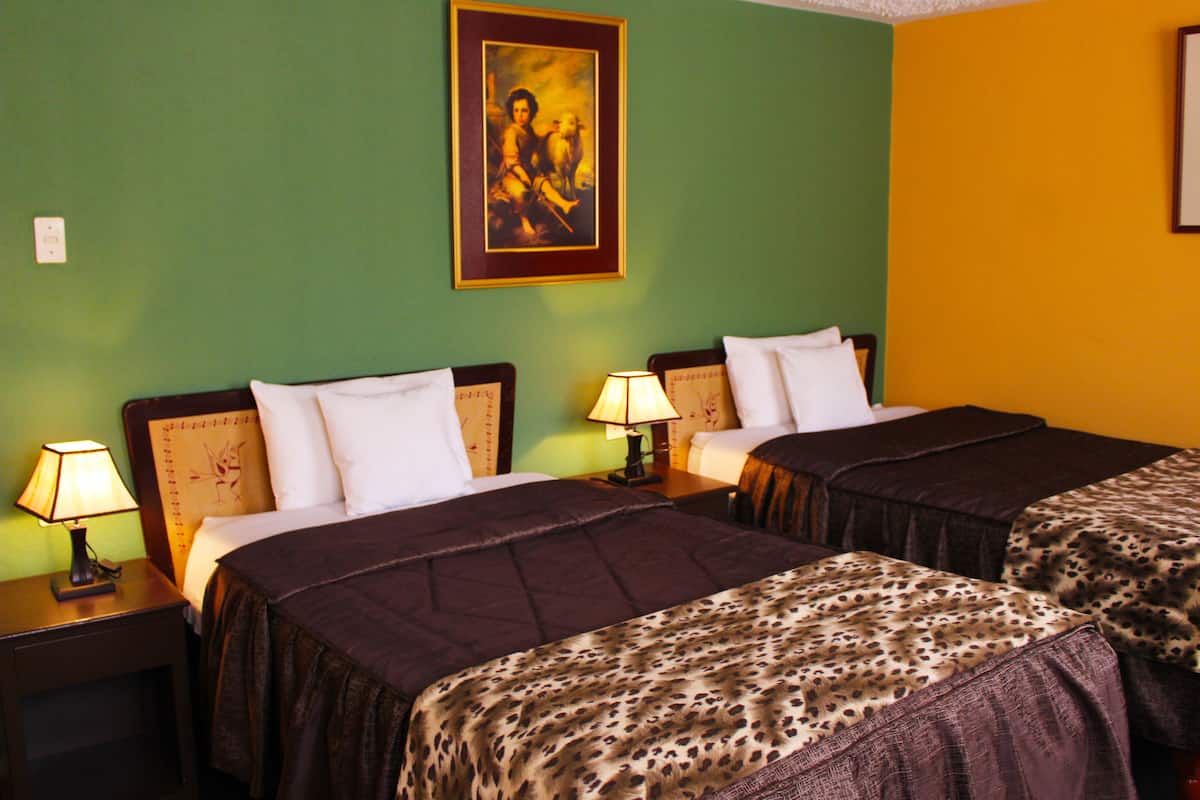 Habitación de hotel con dos camas, cada una con cabecera, mesita de noche con lámpara y un cuadro en la pared.