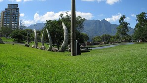 A grassy park with a tall building in the background, a mountain range, and a clear sky. - Hotel Columbus (San José)