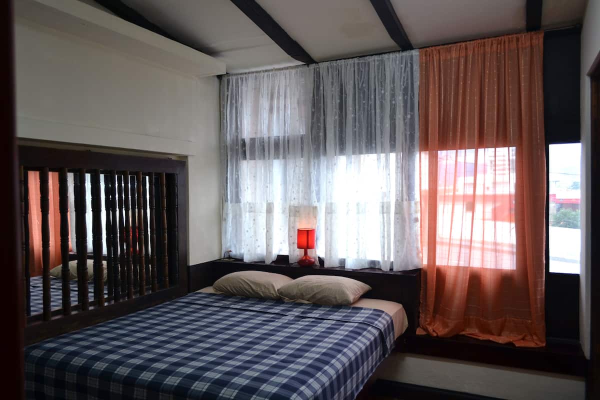 A bedroom with a bed, a window with curtains, a nightstand with a lamp, and a wooden headboard.