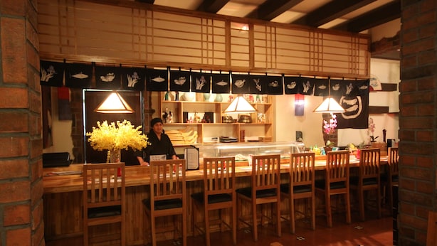 A restaurant with wooden furniture, a counter, and a person serving. - Hotel Columbus (San José)