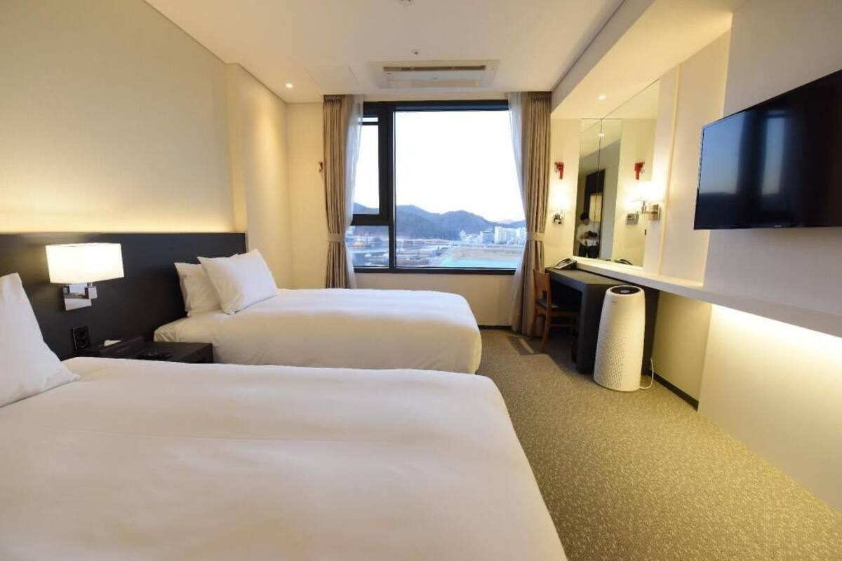 Hotelkamer met twee bedden, een bureau, een stoel, een televisie en een groot raam met uitzicht op de stad.