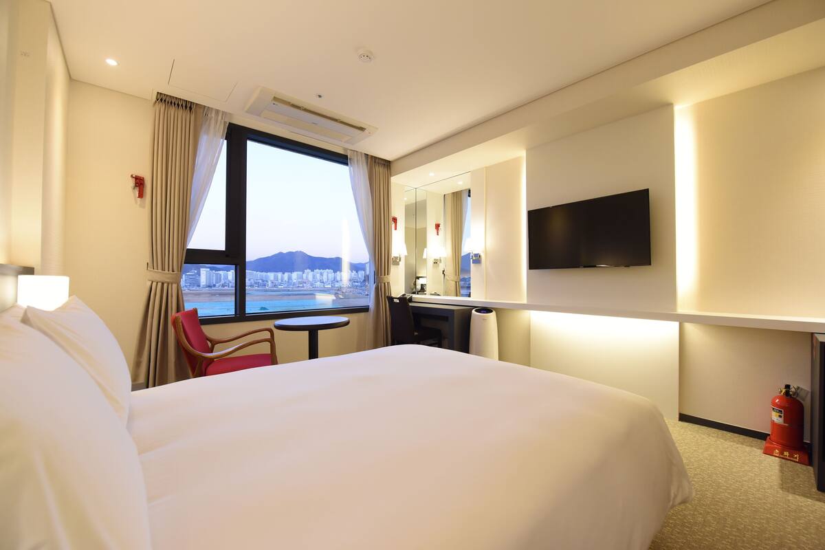 Ein Hotelzimmer mit einem großen Bett, einem Schreibtisch, einem Sessel, einem Fernseher und einem Fenster mit Blick auf die Stadt.