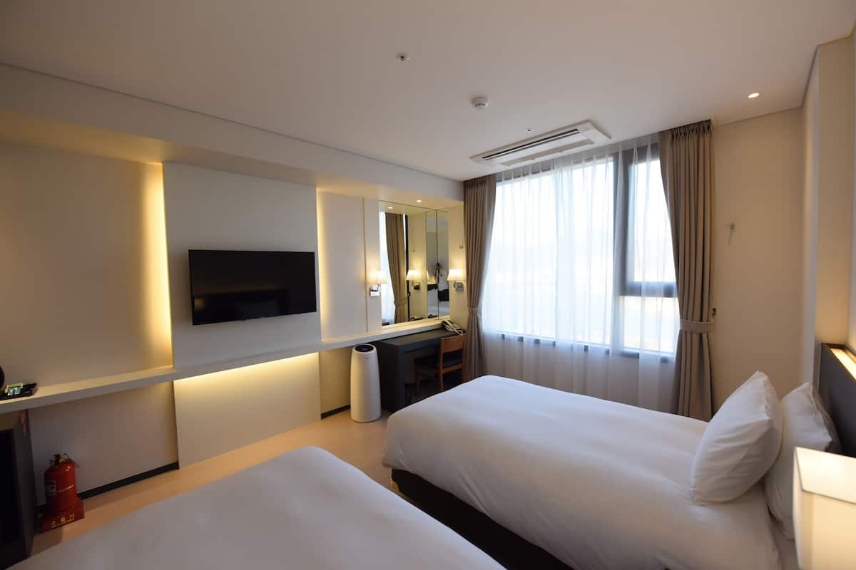 Ein Hotelzimmer mit zwei Betten, einem Fernseher, einem Schreibtisch mit Stuhl, einem Spiegel und einem Fenster mit Vorhängen.