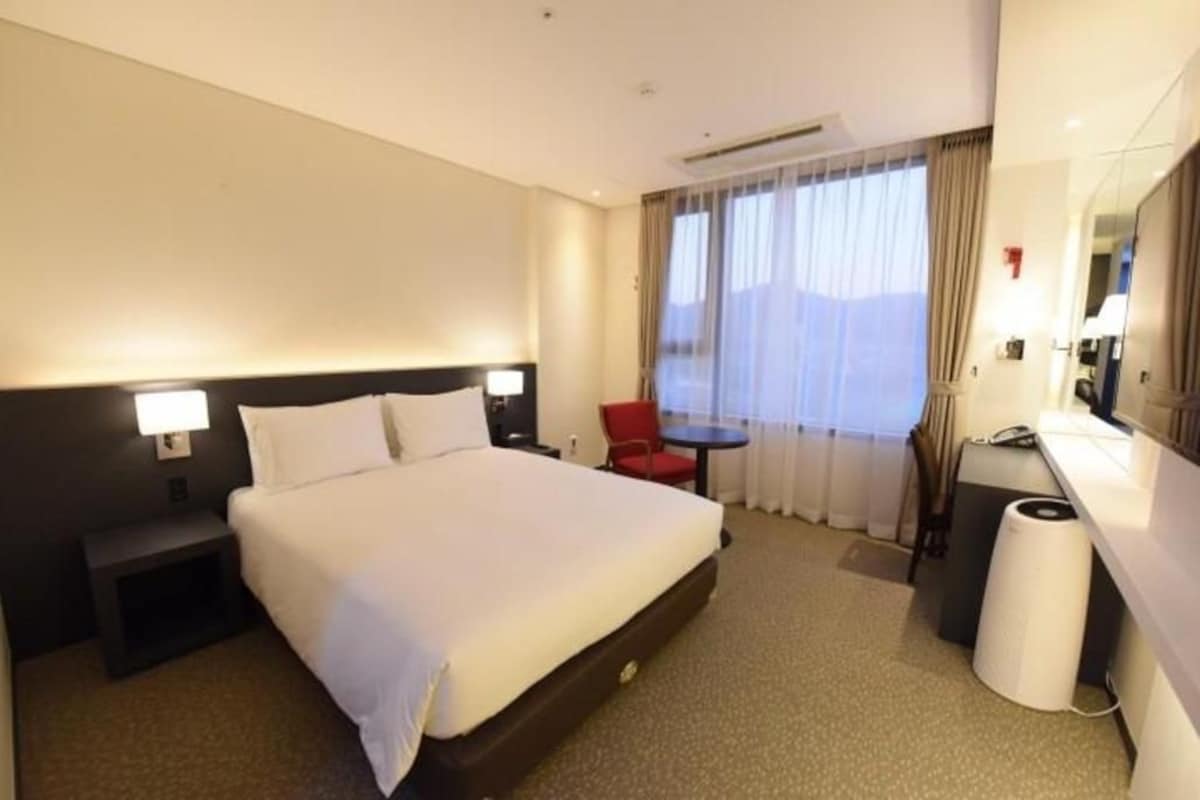 Une chambre d’hôtel comprenant un lit, une table de chevet, un bureau, une chaise et une grande fenêtre avec des rideaux.