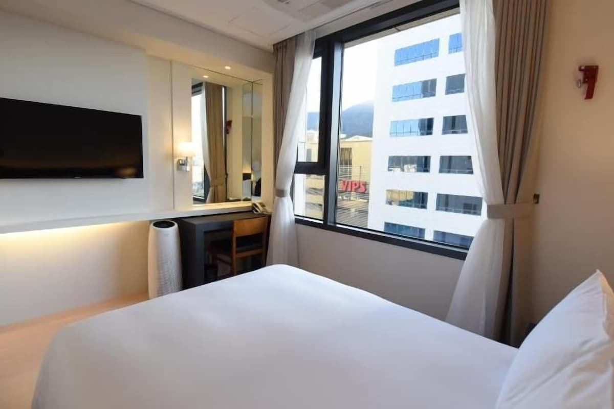 Une chambre d’hôtel équipée d’un lit, d’un téléviseur à écran plat, d’un bureau et d’une fenêtre donnant sur la ville.