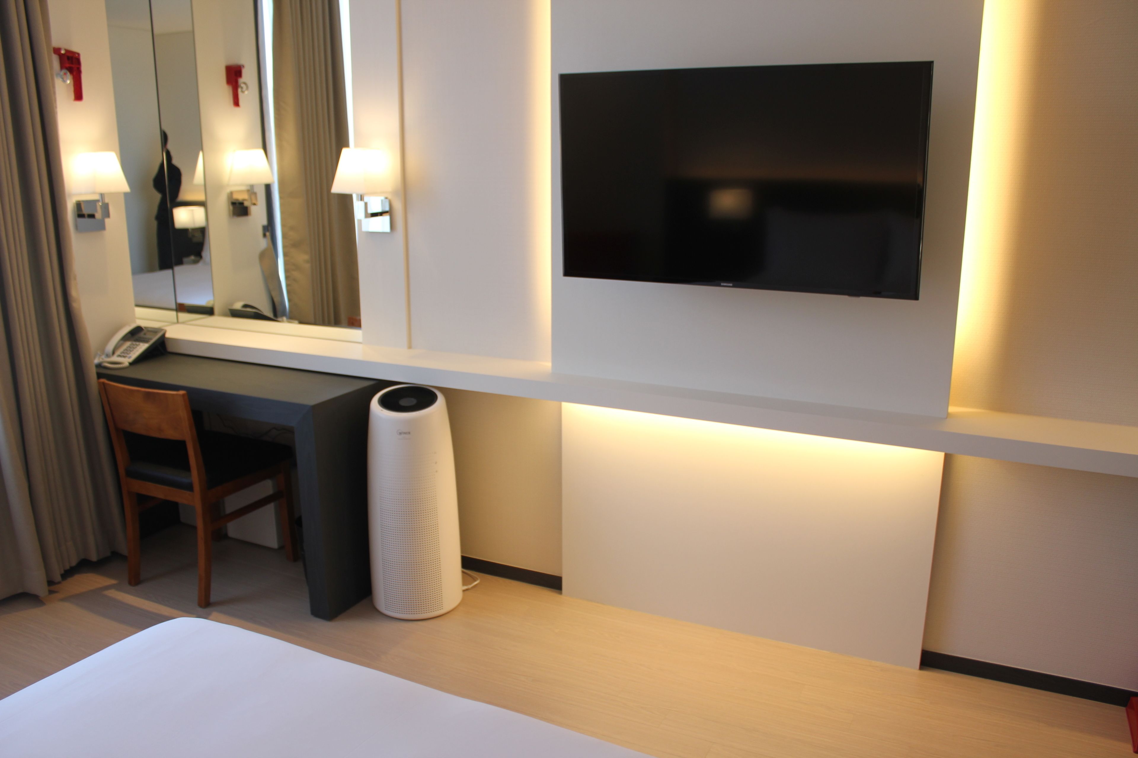 A hotel room with a bed, a desk, a chair, a telephone, a trash bin, a mirror, and a flat-screen TV mounted on the wall.