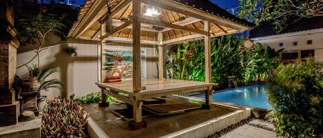 Un quiosco de madera con cerramiento de vidrio, piscina y plantas tropicales.