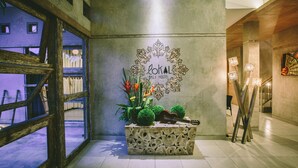 Área de lobby com um banco de madeira, parede decorativa com o texto "Lokal Bali Hostel" e um pequeno vaso com plantas e um violão.