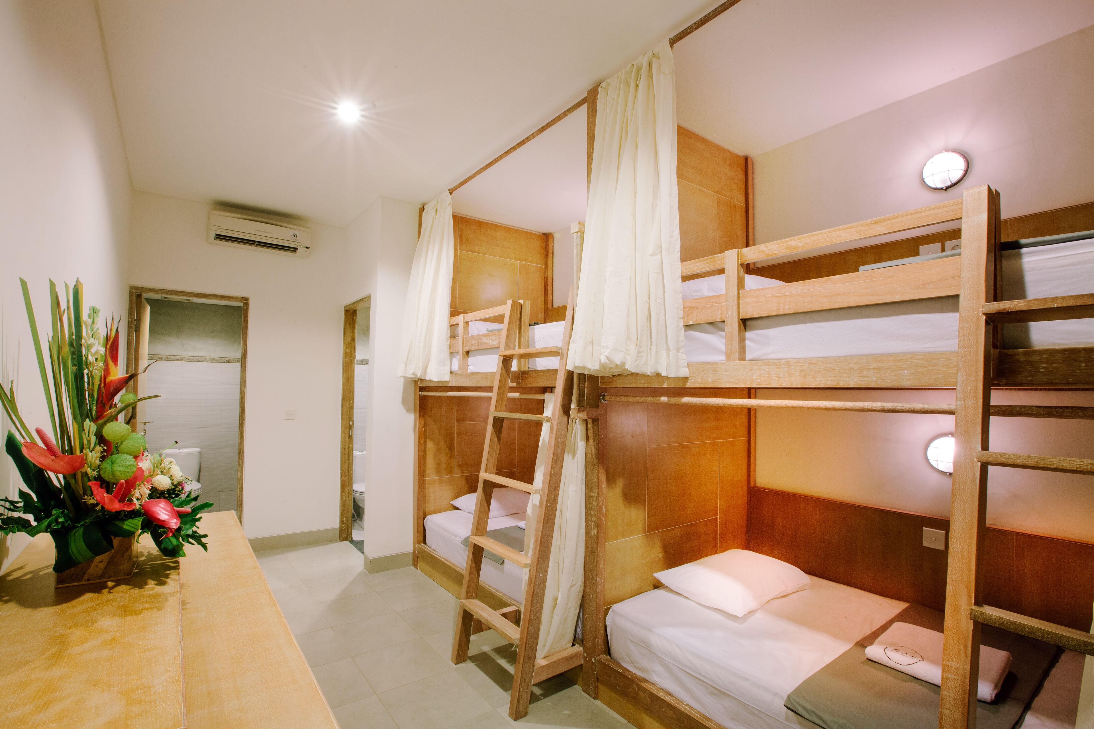 Une chambre avec un lit superposé, des montants en bois, une literie blanche et un bureau en bois agrémenté d’un vase de fleurs.