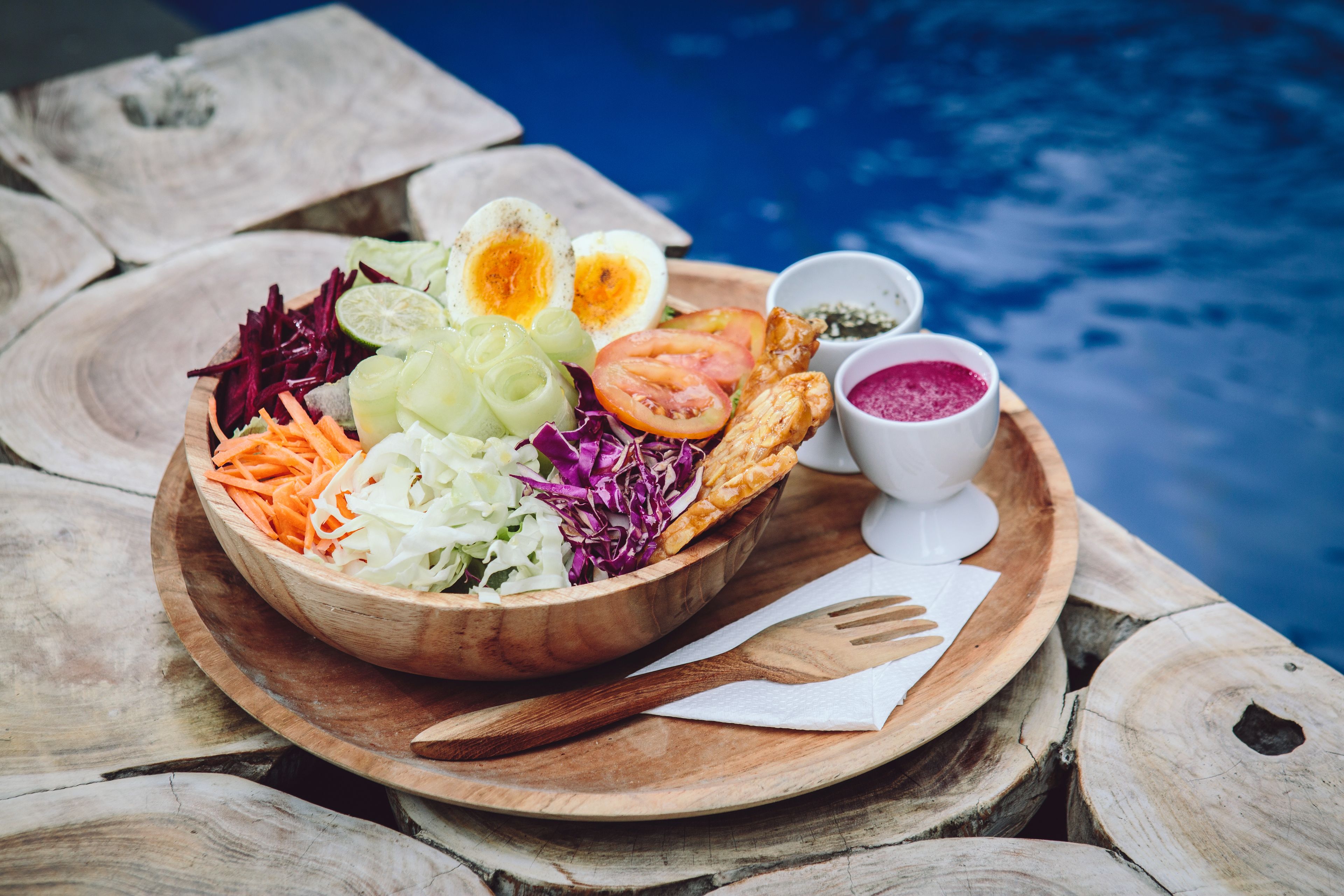 A wooden bowl filled with salad, eggs, and bread, placed on a wooden table by a pool.