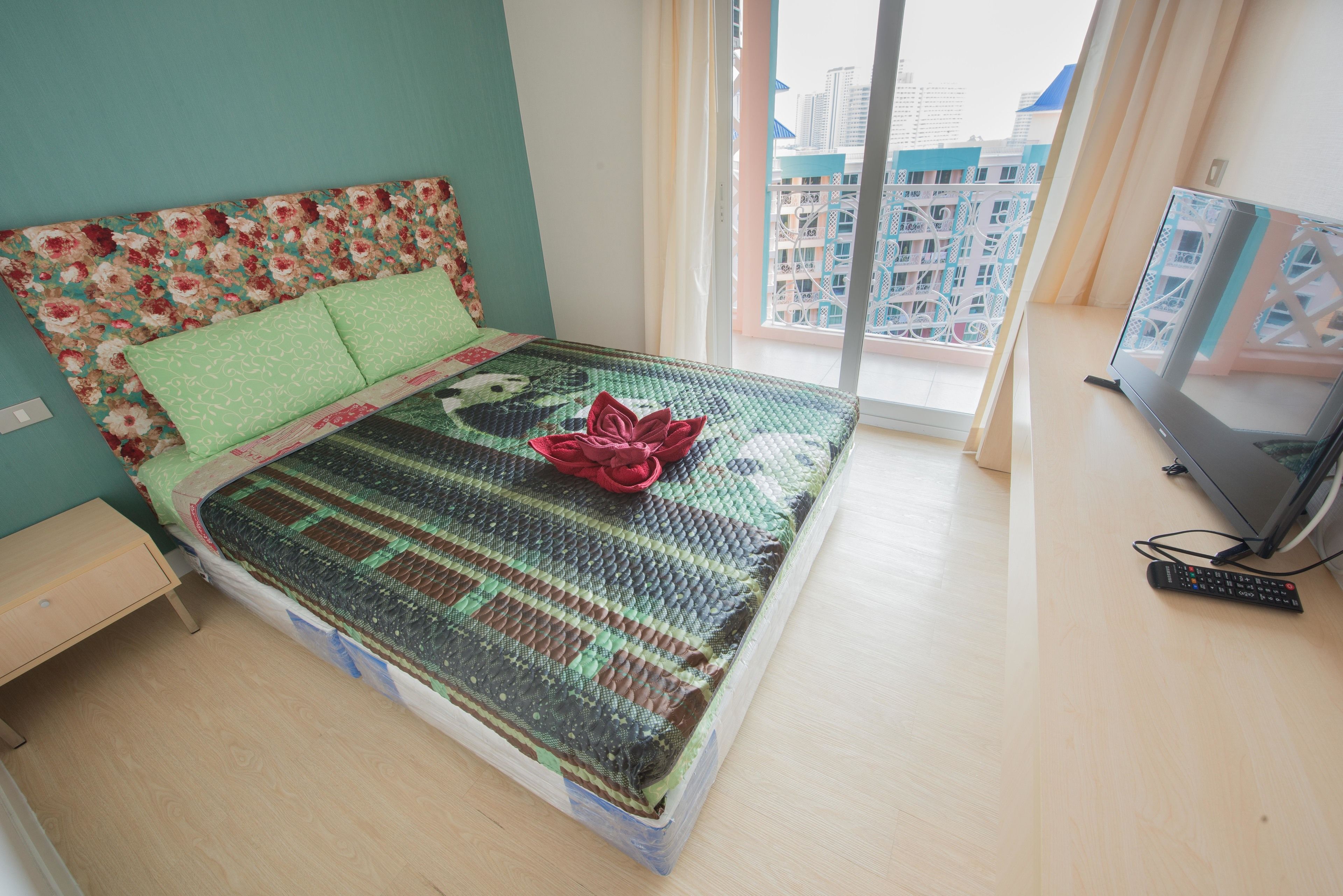 A neatly made bed with a floral headboard, a green patterned bedspread, and a red decorative pillow. A remote control is on the wooden bedside table.