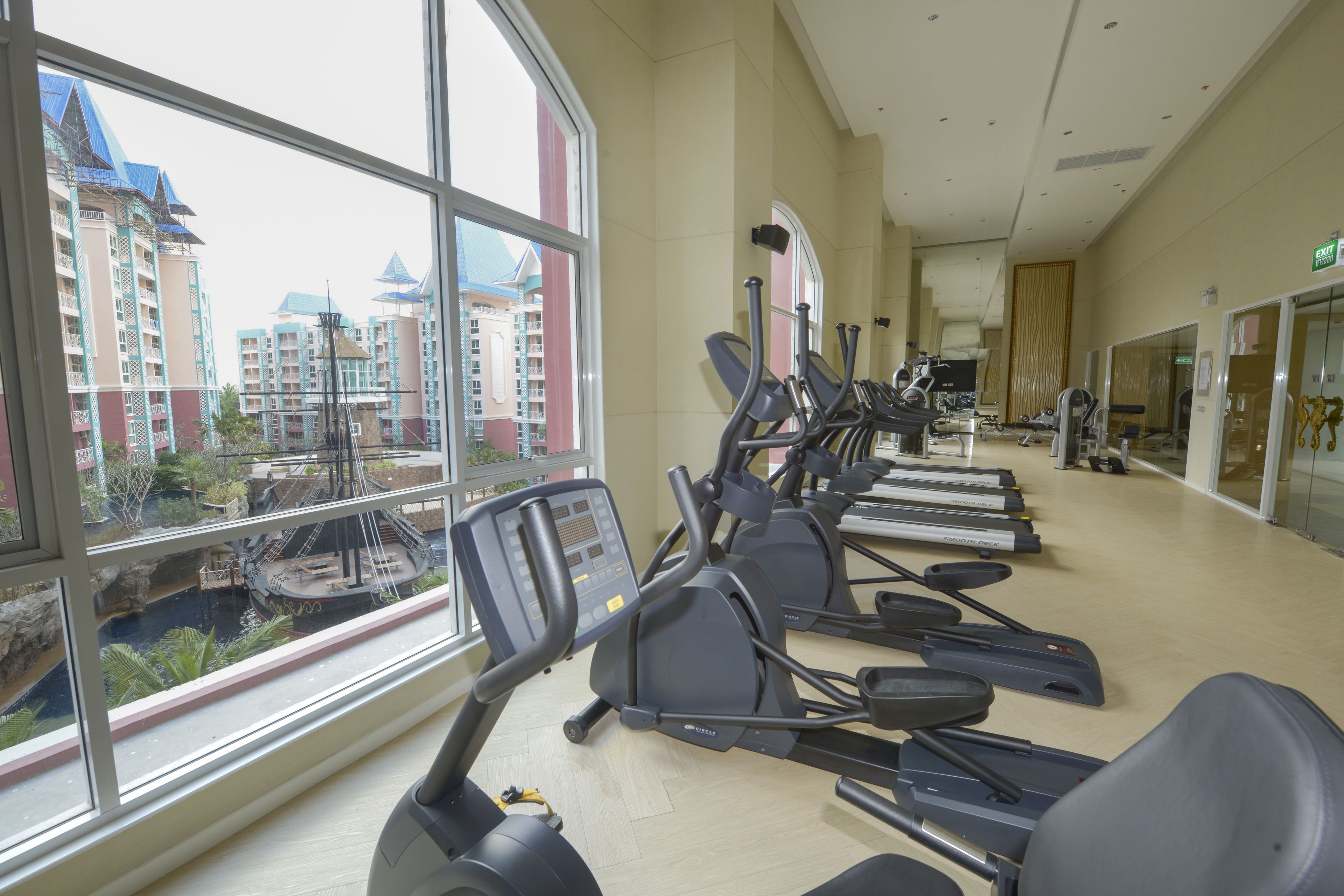 A gym with treadmills and exercise equipment, large windows with a view of buildings, and a wooden floor.
