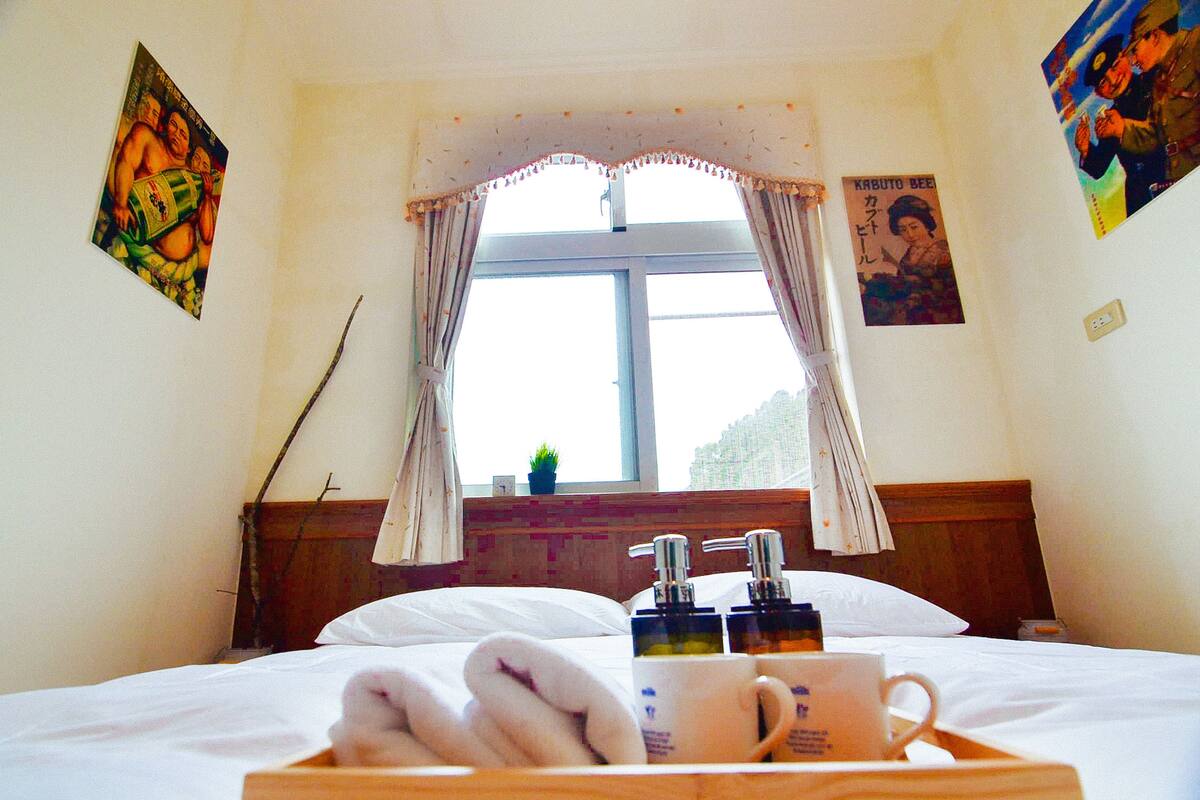 Habitación de hotel con cama, ventana con cortinas, mesita de noche con artículos de aseo y decoración en las paredes.