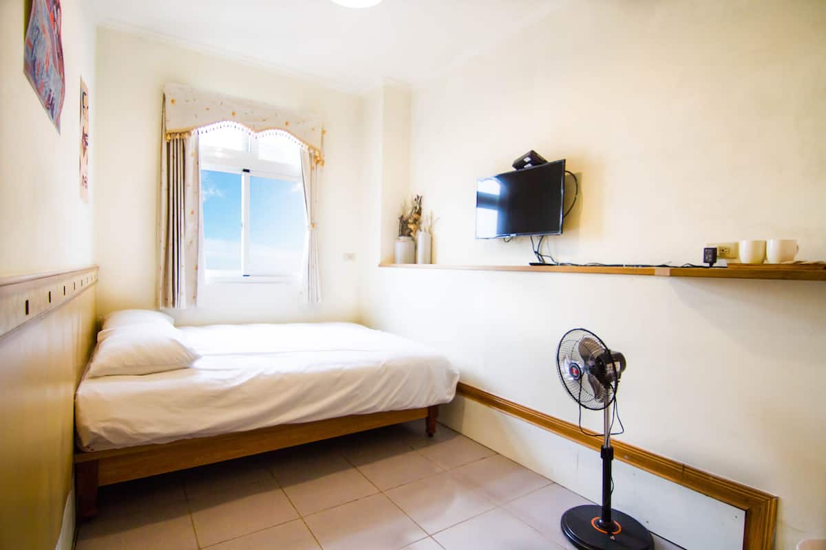 Un dormitorio con una cama, televisión, ventilador y ventana con cortinas.