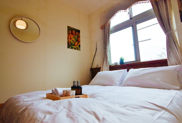 Uma cama bem arrumada com lençóis brancos, uma mesinha de cabeceira com um porta-objetos e uma janela com cortinas.