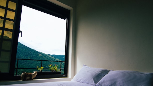Uma cama com roupa de cama branca, uma janela com vista para uma montanha e uma pequena planta em vaso no parapeito.
