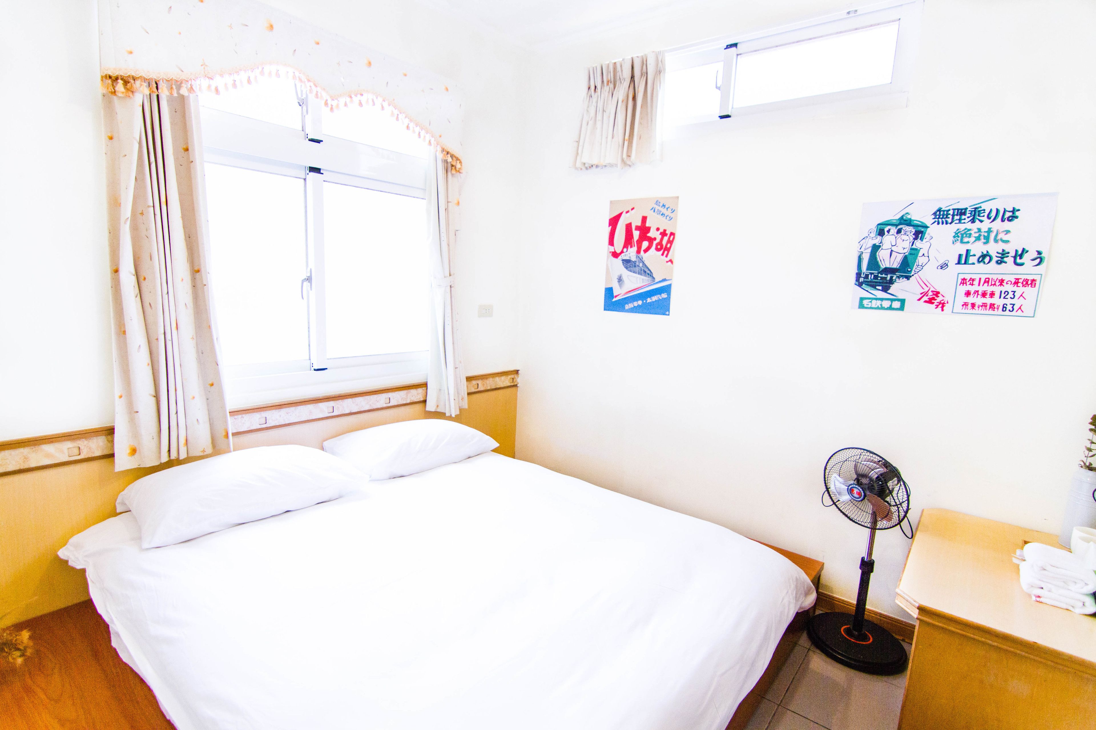A hotel room with a bed, a standing fan, a window with curtains, and a wall with posters.