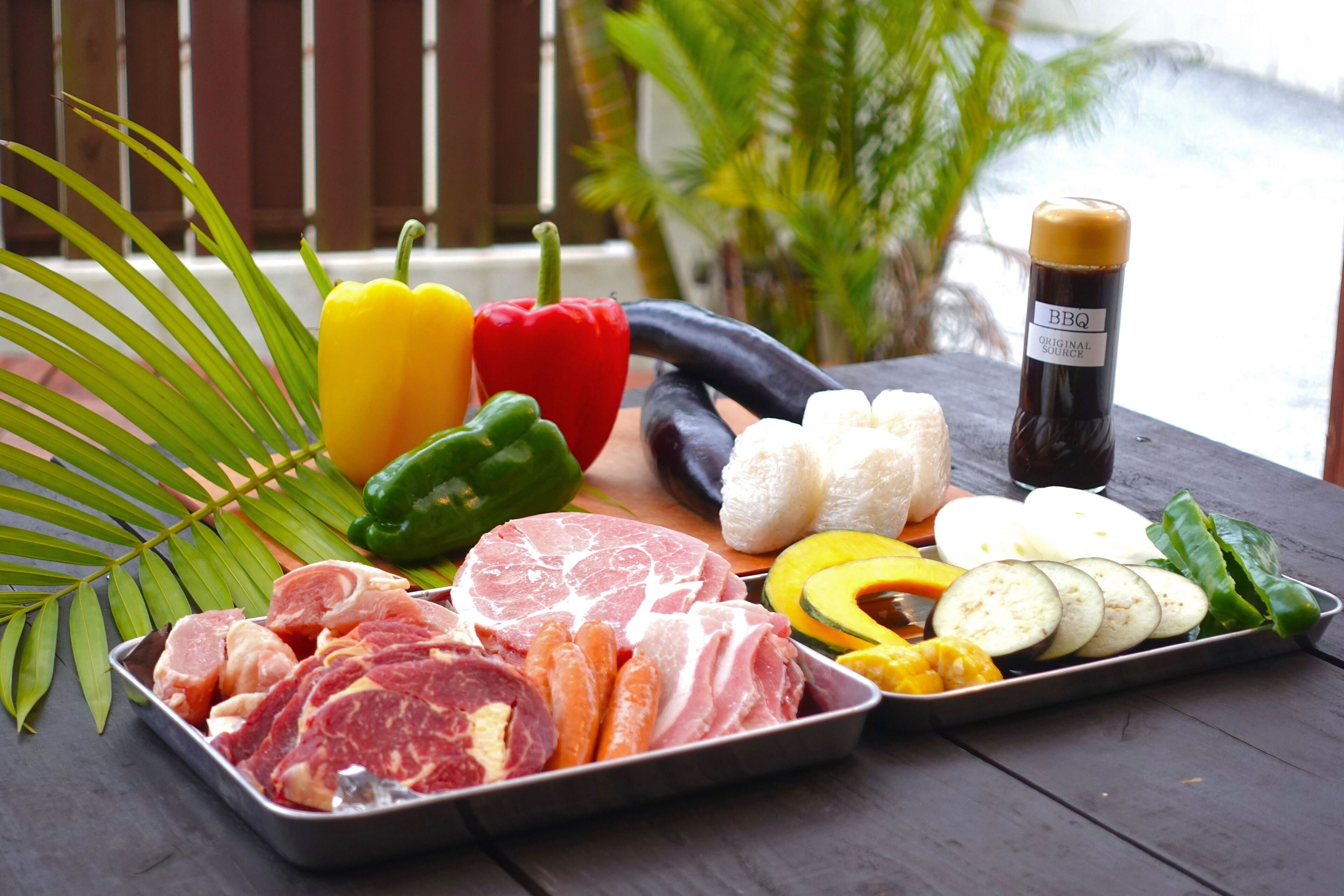 Diverse rauw vlees, groenten en een fles barbecuesaus op een houten tafel buiten.