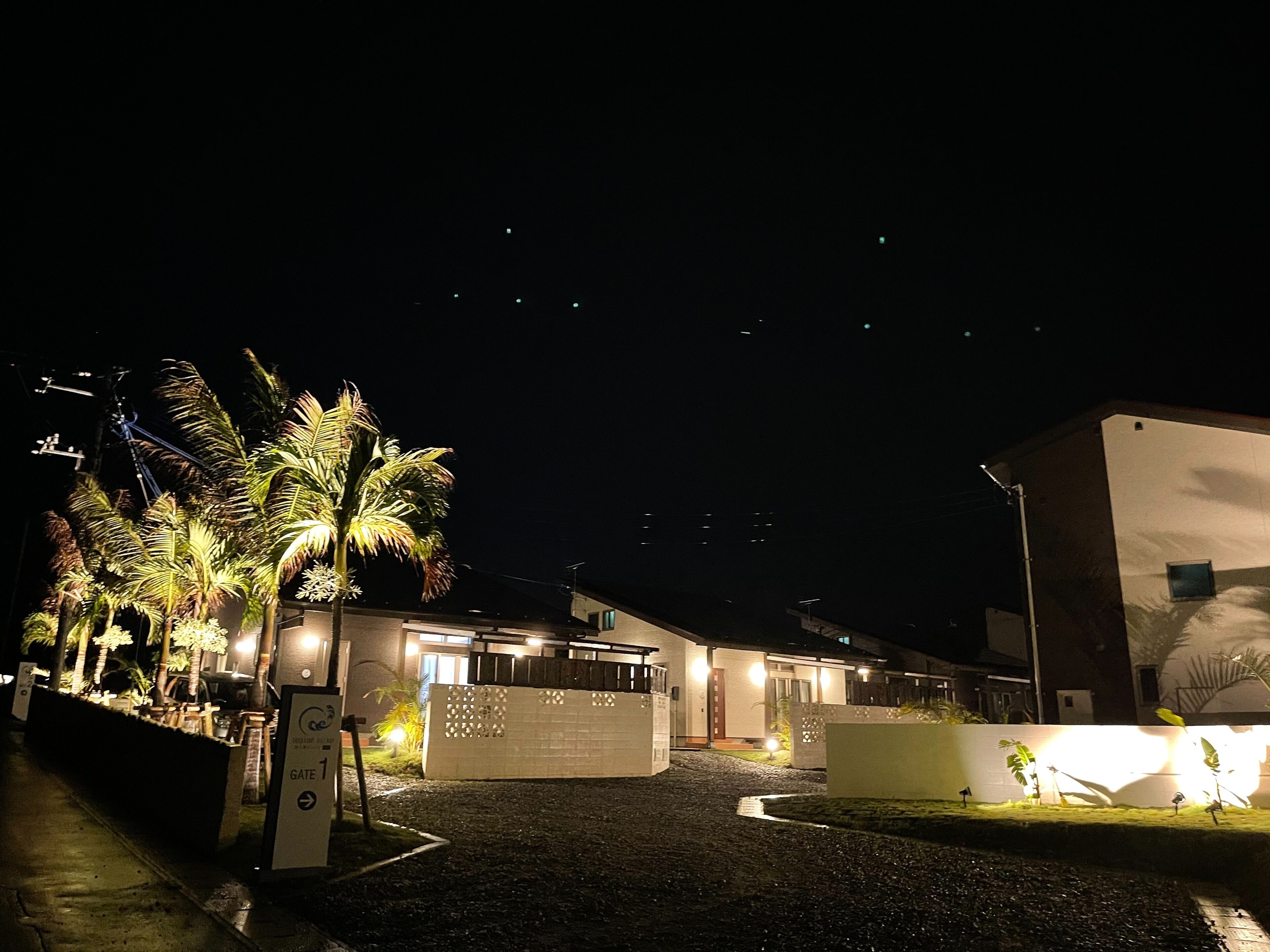 A nighttime view of a building with a paved entrance area, palm trees, and a sign indicating a gate.