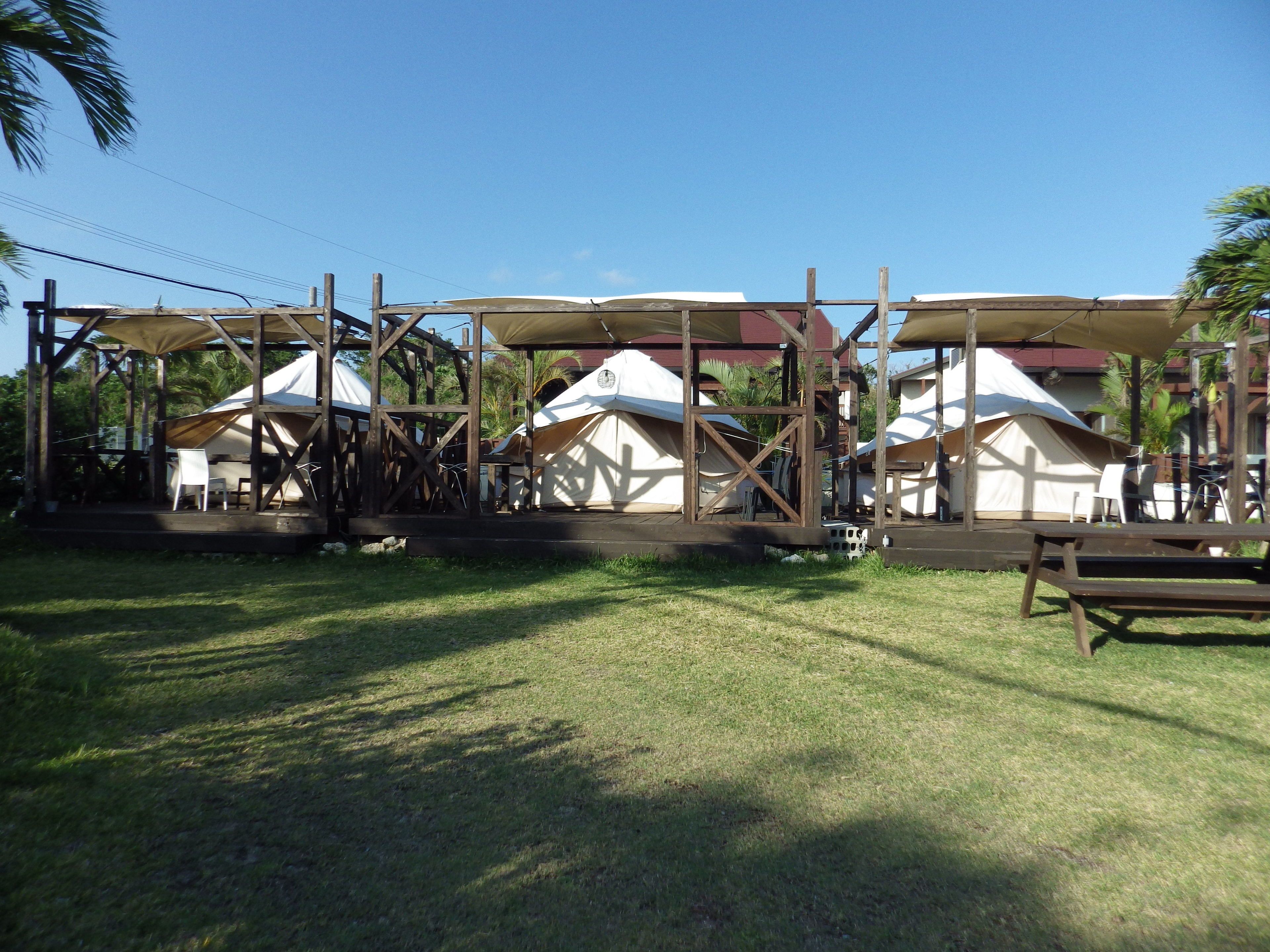 A series of yurts with wooden frames and canvas roofs, set on a grassy area with a wooden bench and tables.