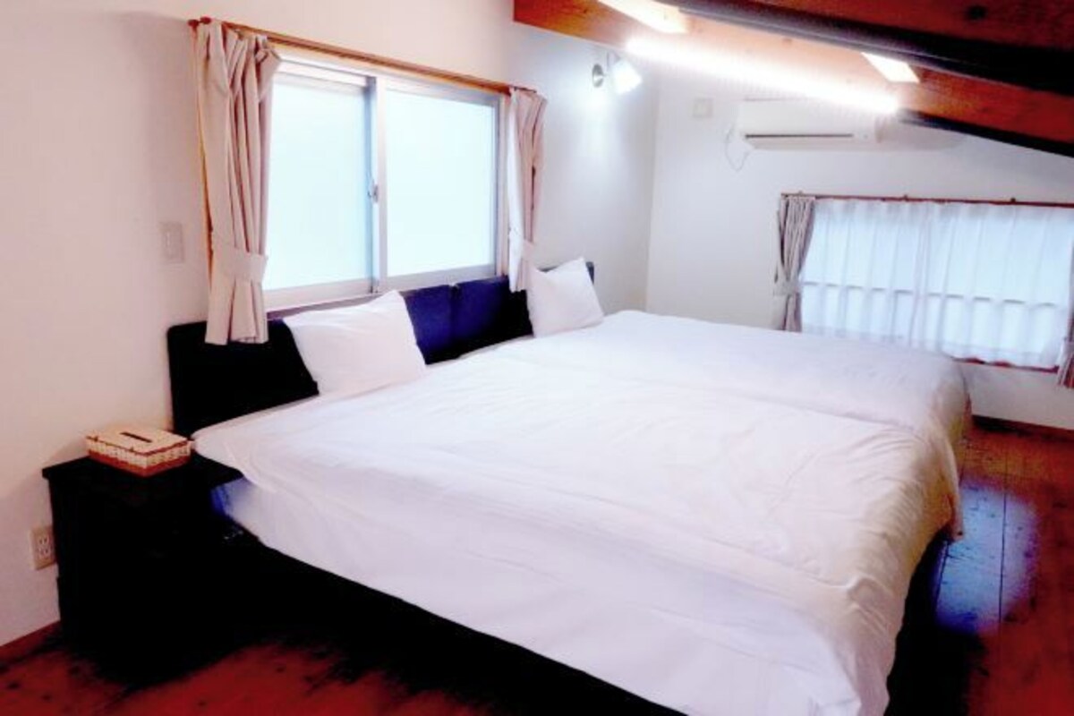A bedroom with a large bed, a nightstand, a window with curtains, and an air conditioning unit.
