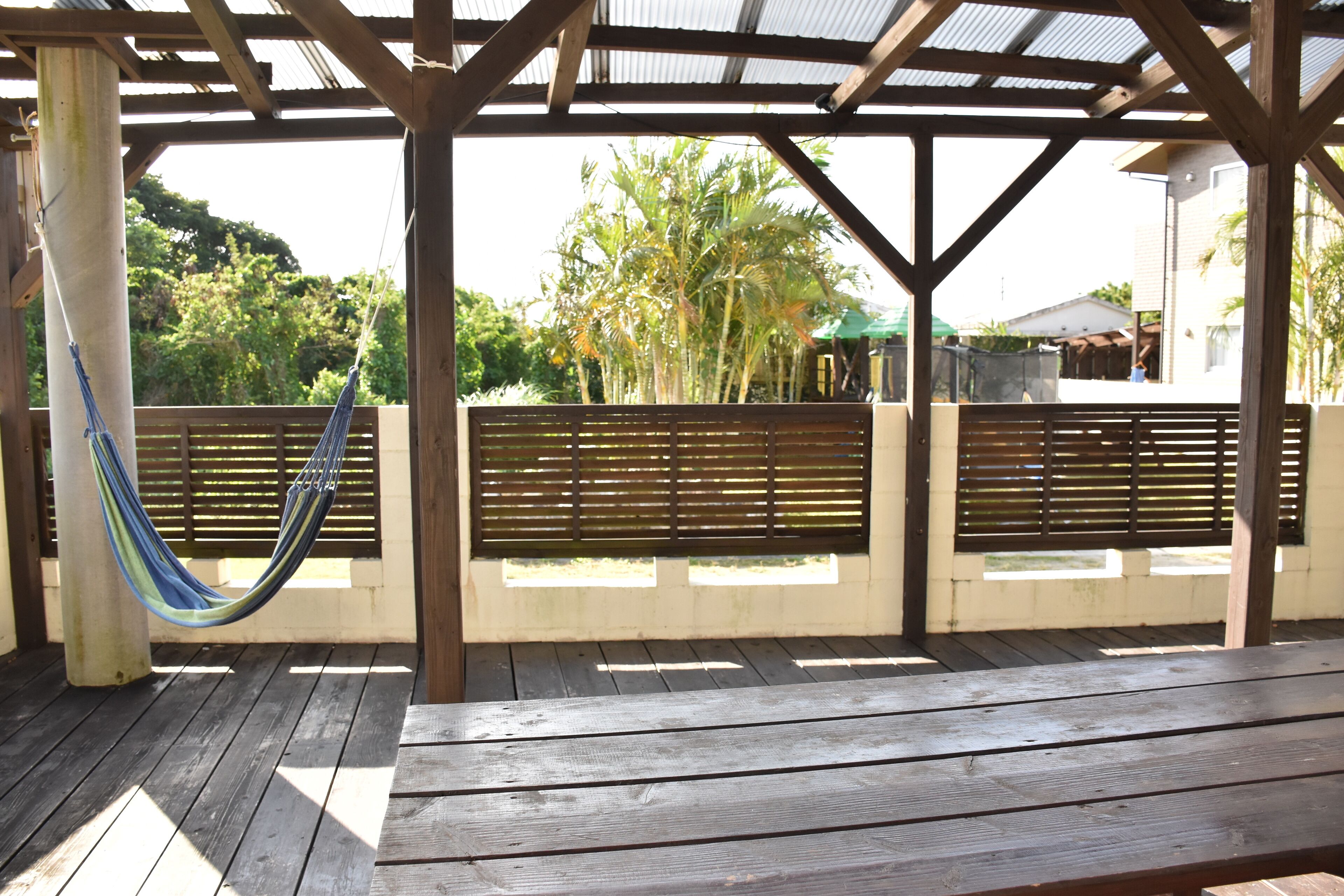 Une terrasse en bois avec un hamac, des bancs en bois et un espace abrité offrant une vue sur la verdure et les bâtiments en arrière-plan.