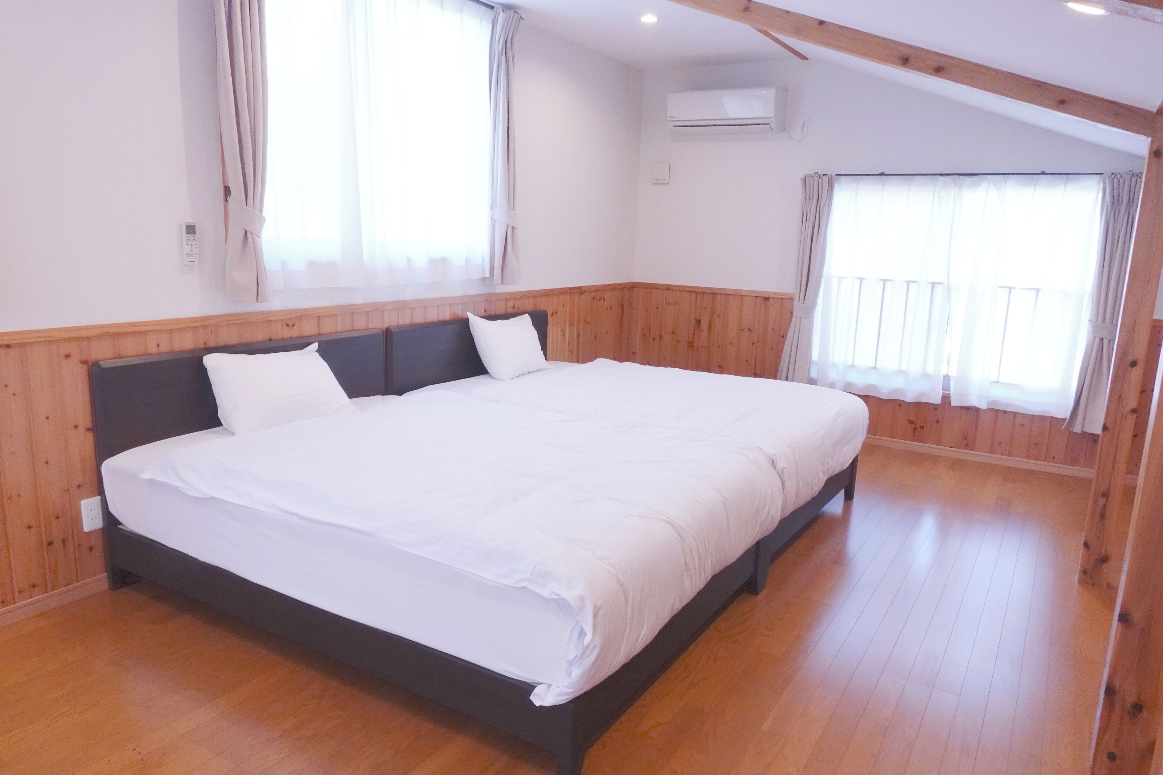 Een groot bed in een kamer met houten lambrisering, een raam met gordijnen en een airconditioning.
