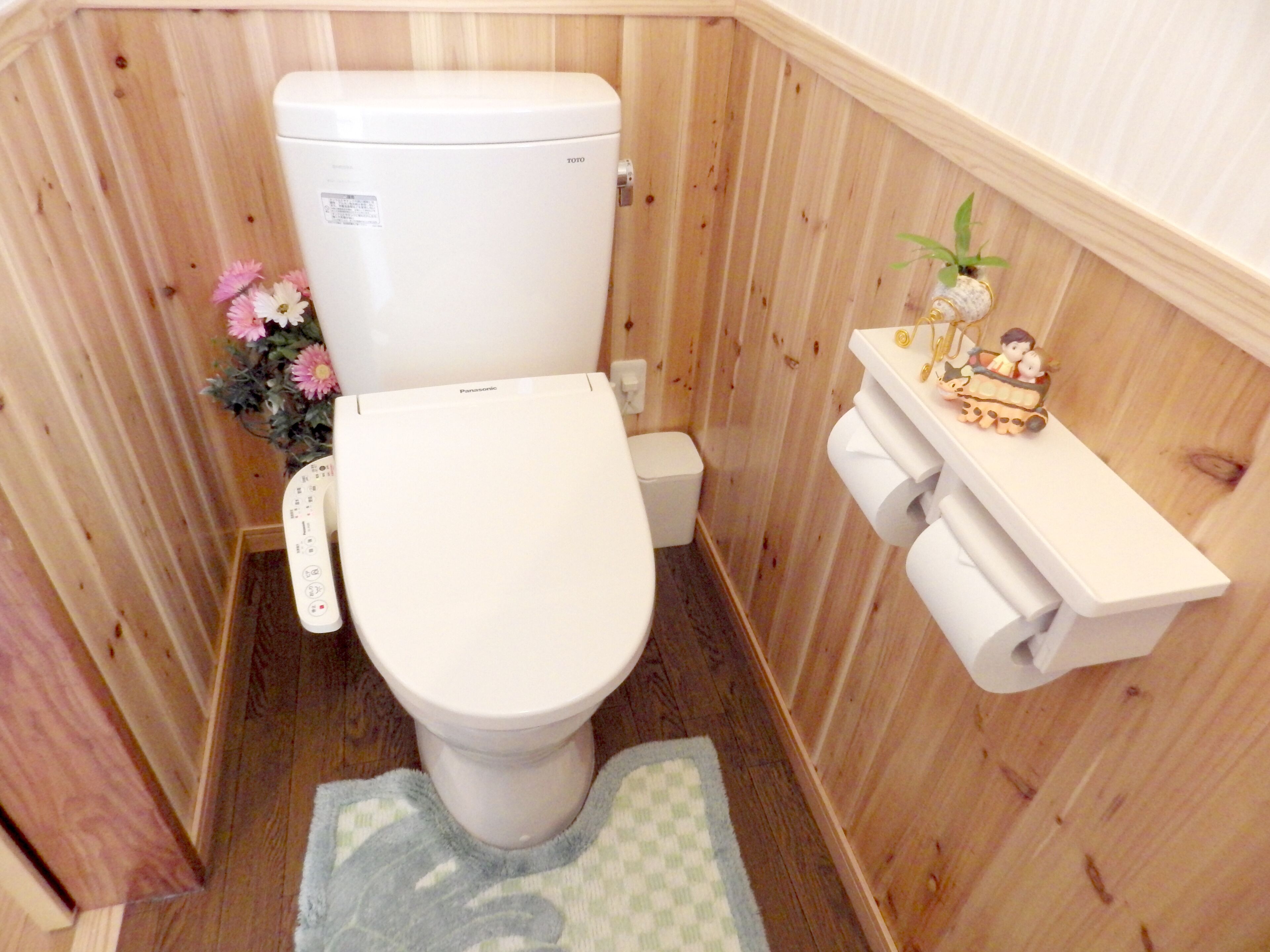 A compact wooden bathroom with a white toilet, a small shelf with decorative items, and a floral arrangement.