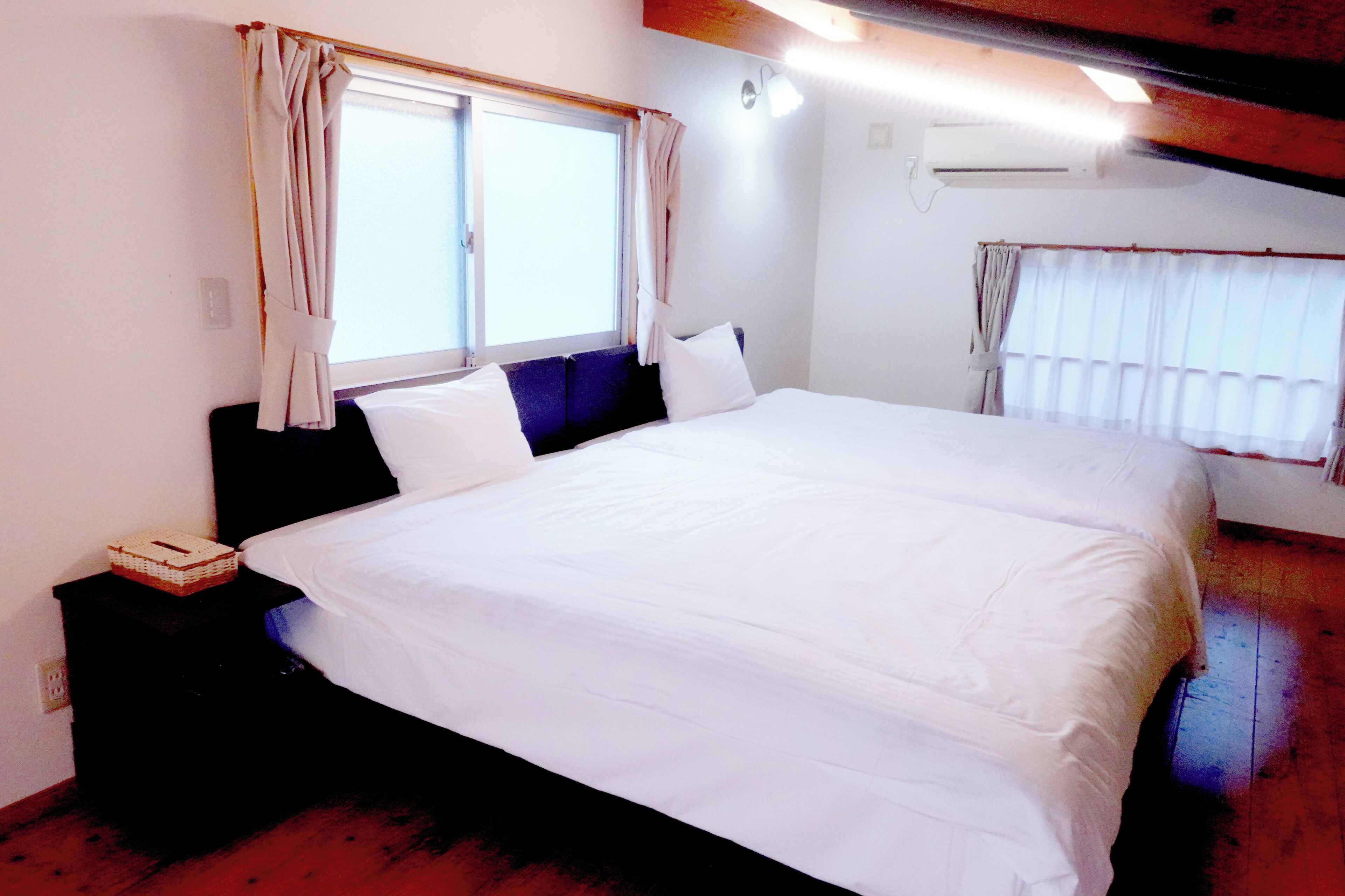 A bedroom with a large bed, a nightstand, an air conditioning unit, and a window with curtains.