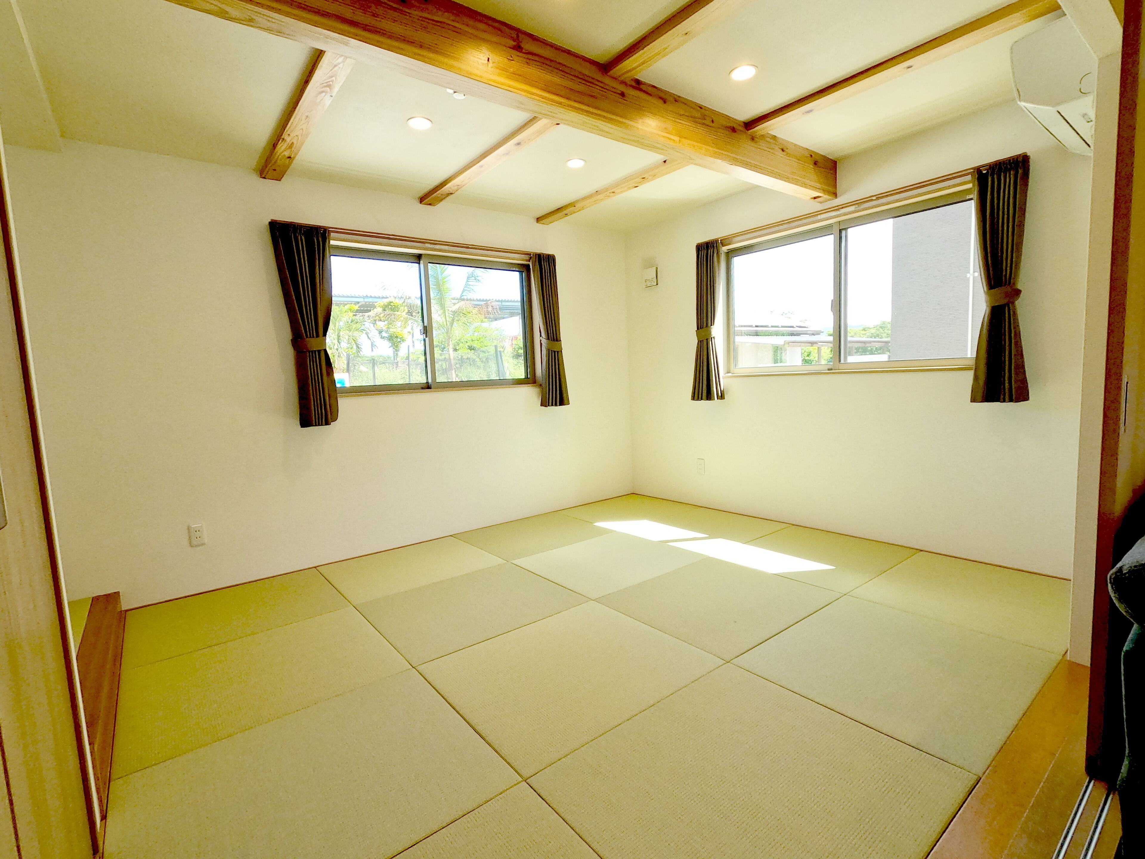 Una habitación con suelo de tatami, vigas de madera y dos ventanas con cortinas.
