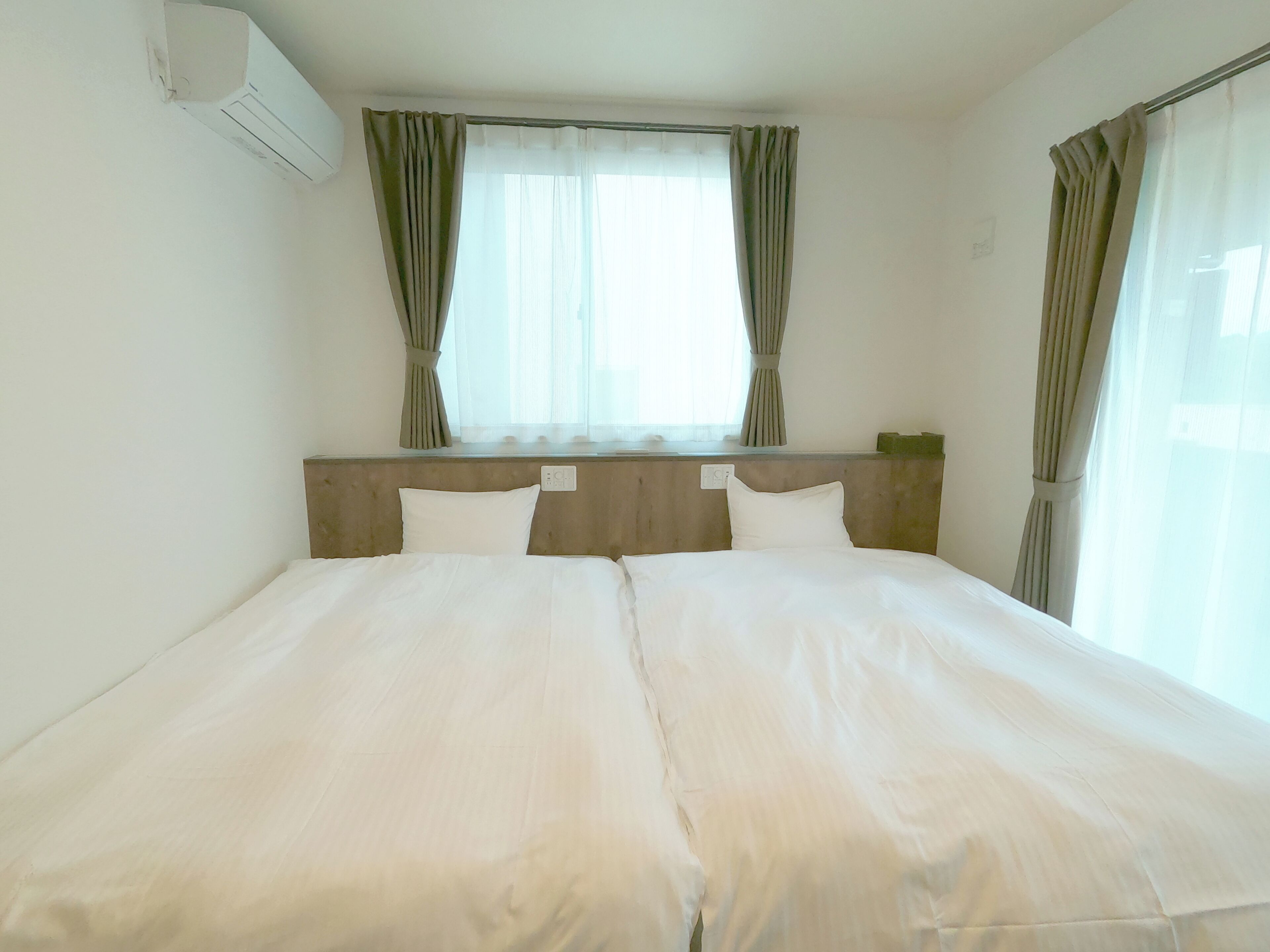 A double bed with white linens, a wooden headboard, and a window with curtains.
