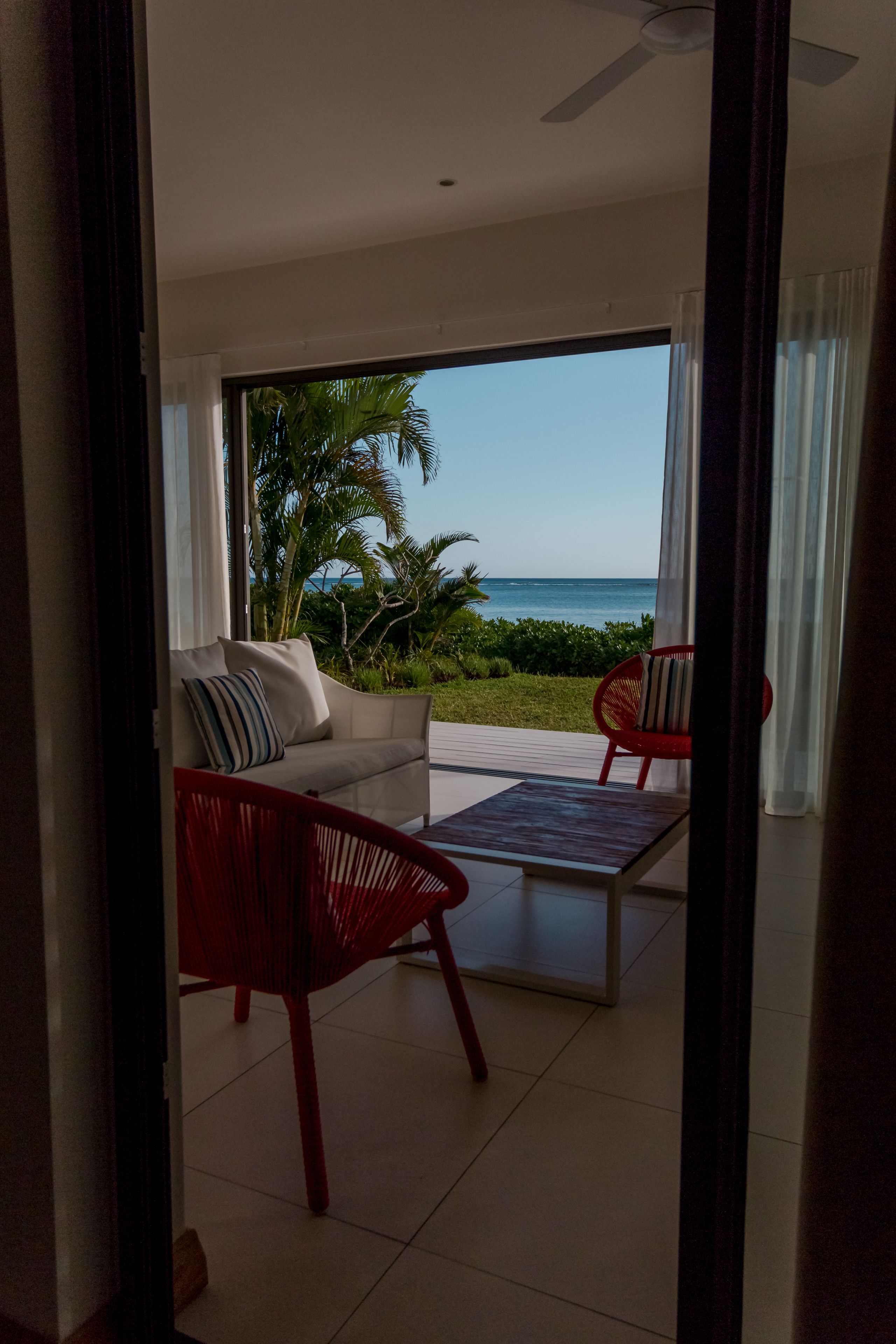 A modern living room with a sofa and two red chairs, a glass coffee table, and a view of the ocean through large windows.