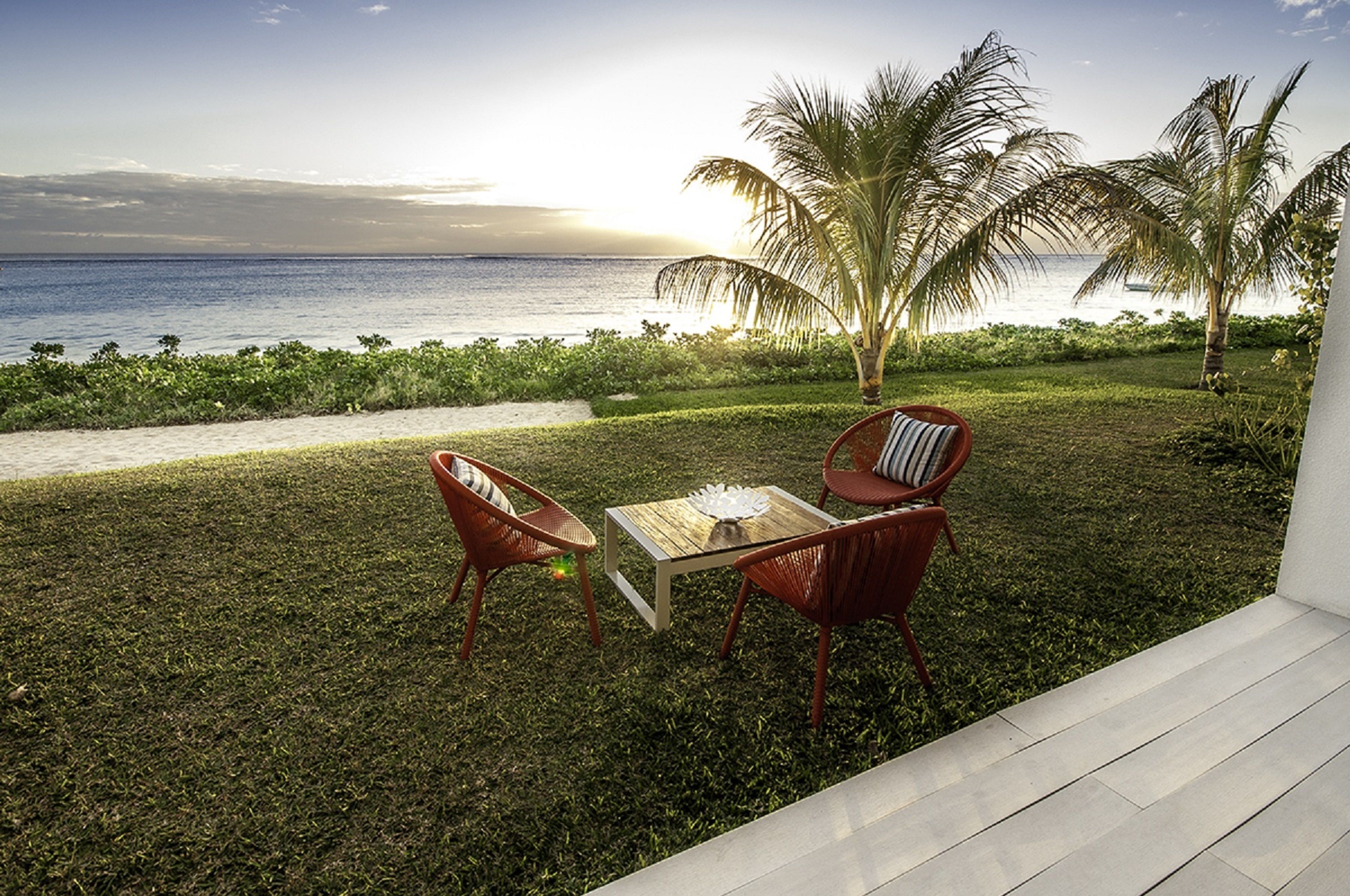 Une terrasse avec deux chaises rouges et une table, donnant sur une plage bordée de palmiers et sous un ciel dégagé.