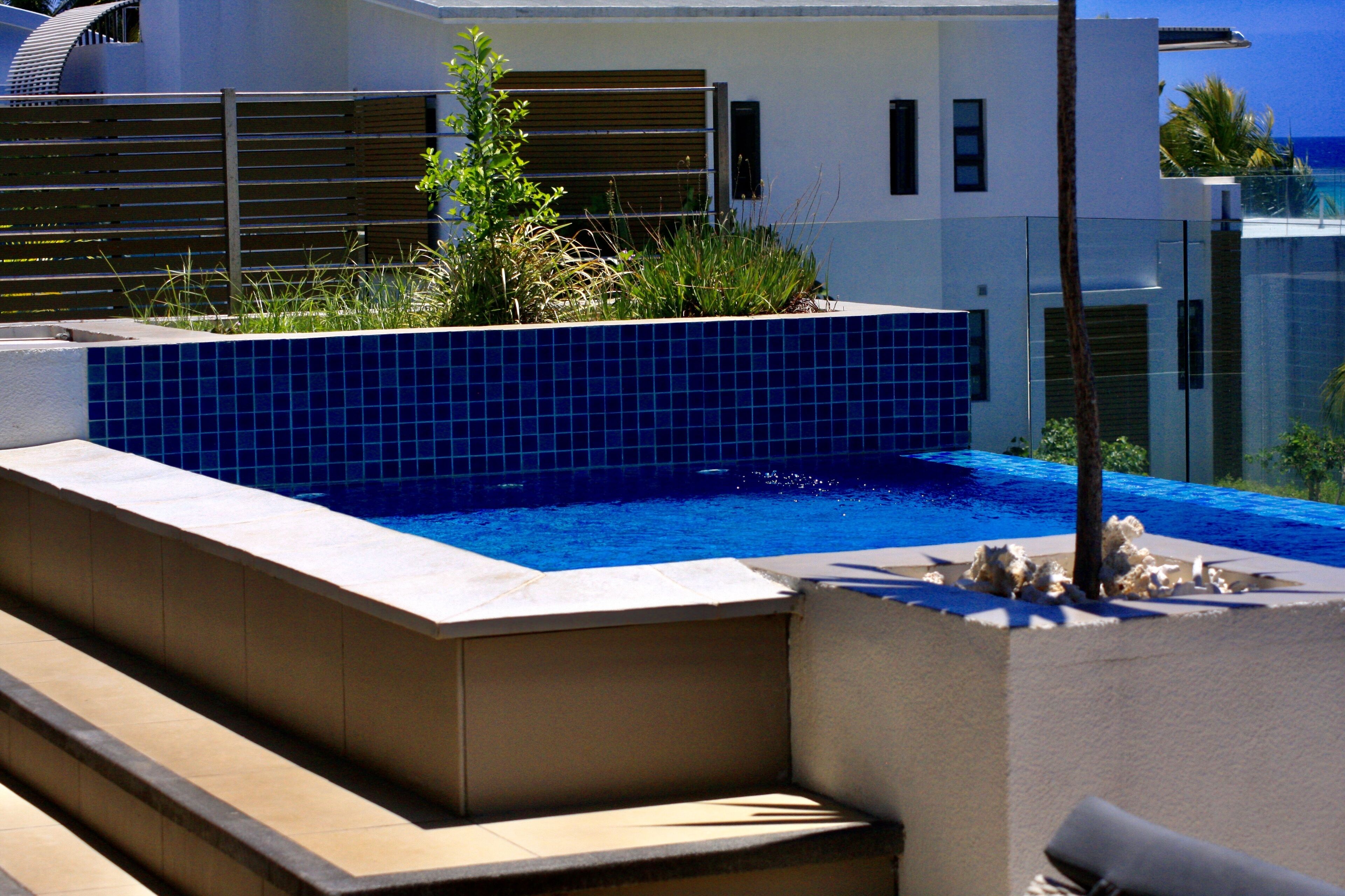 Una piscina esterna moderna con bordi piastrellati di blu, circondata da un muretto basso e gradini che conducono all'acqua.
