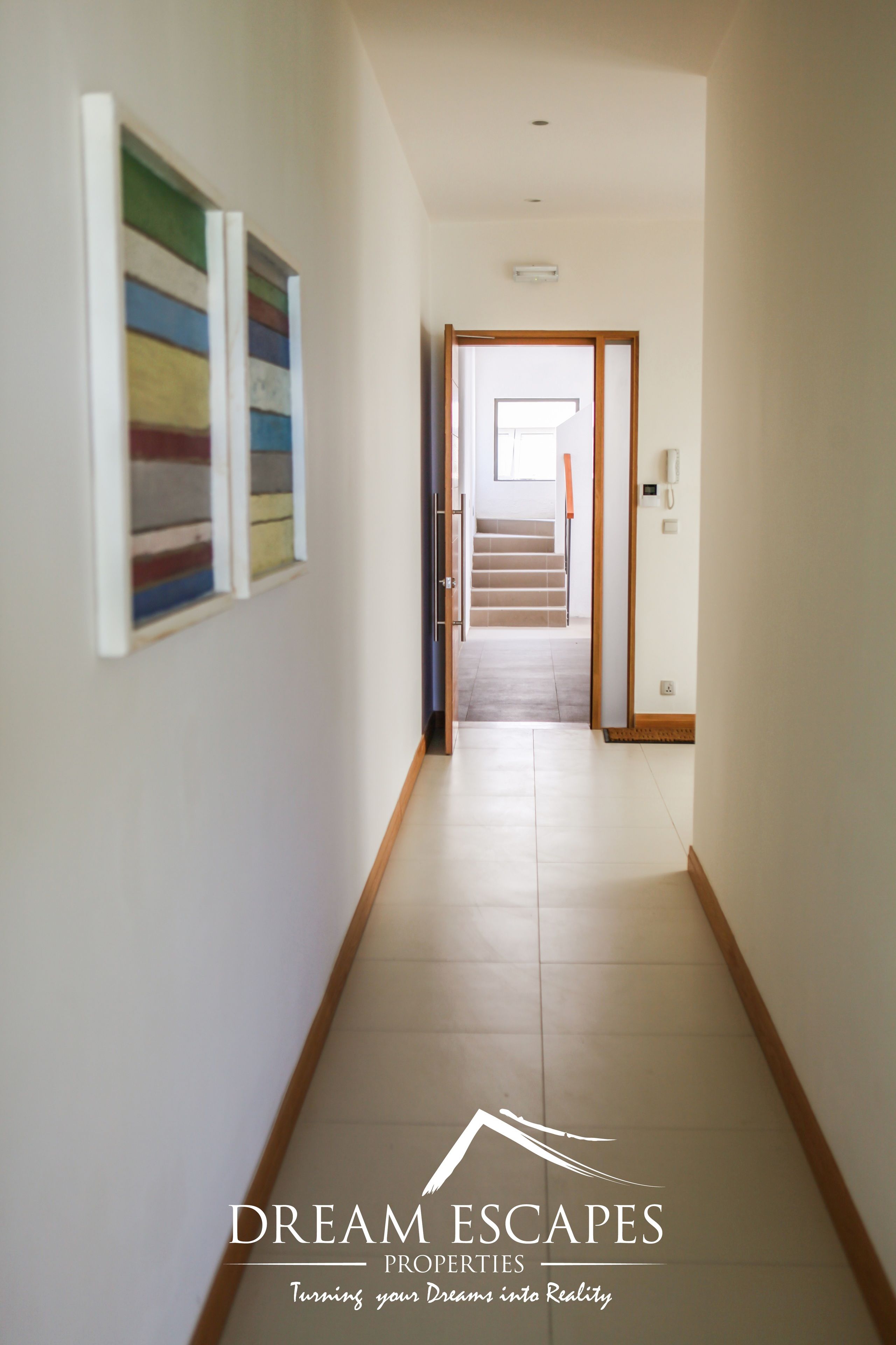 A hallway with a staircase leading to another room. There are two framed artworks on the wall. The floor is tiled.