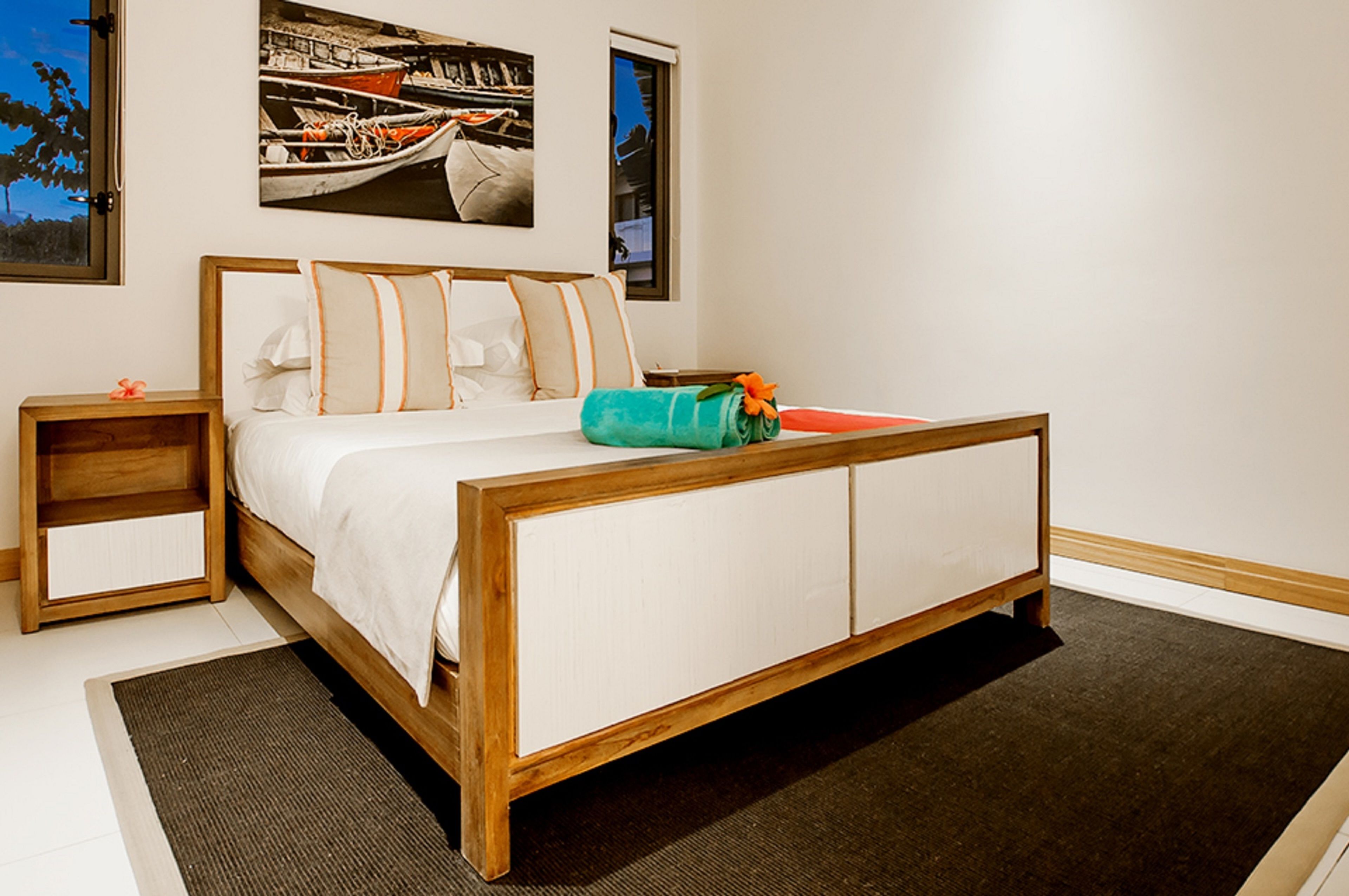 A bedroom with a wooden bed, white bedding, a small nightstand, and a painting of boats on the wall.